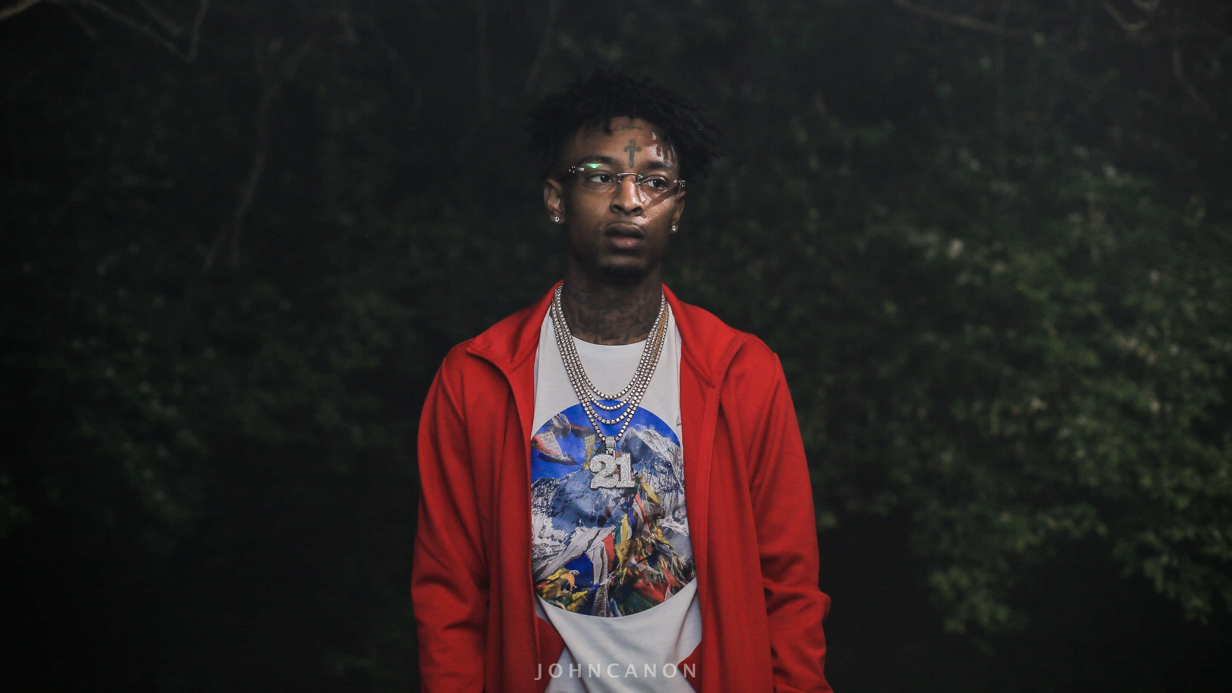 21 Savage 舞台演出 壁紙 — 一個穿著紅色連帽衫和圖案T恤的年輕人，在夜晚的黑暗森林中自信地站著，眼睛發光。