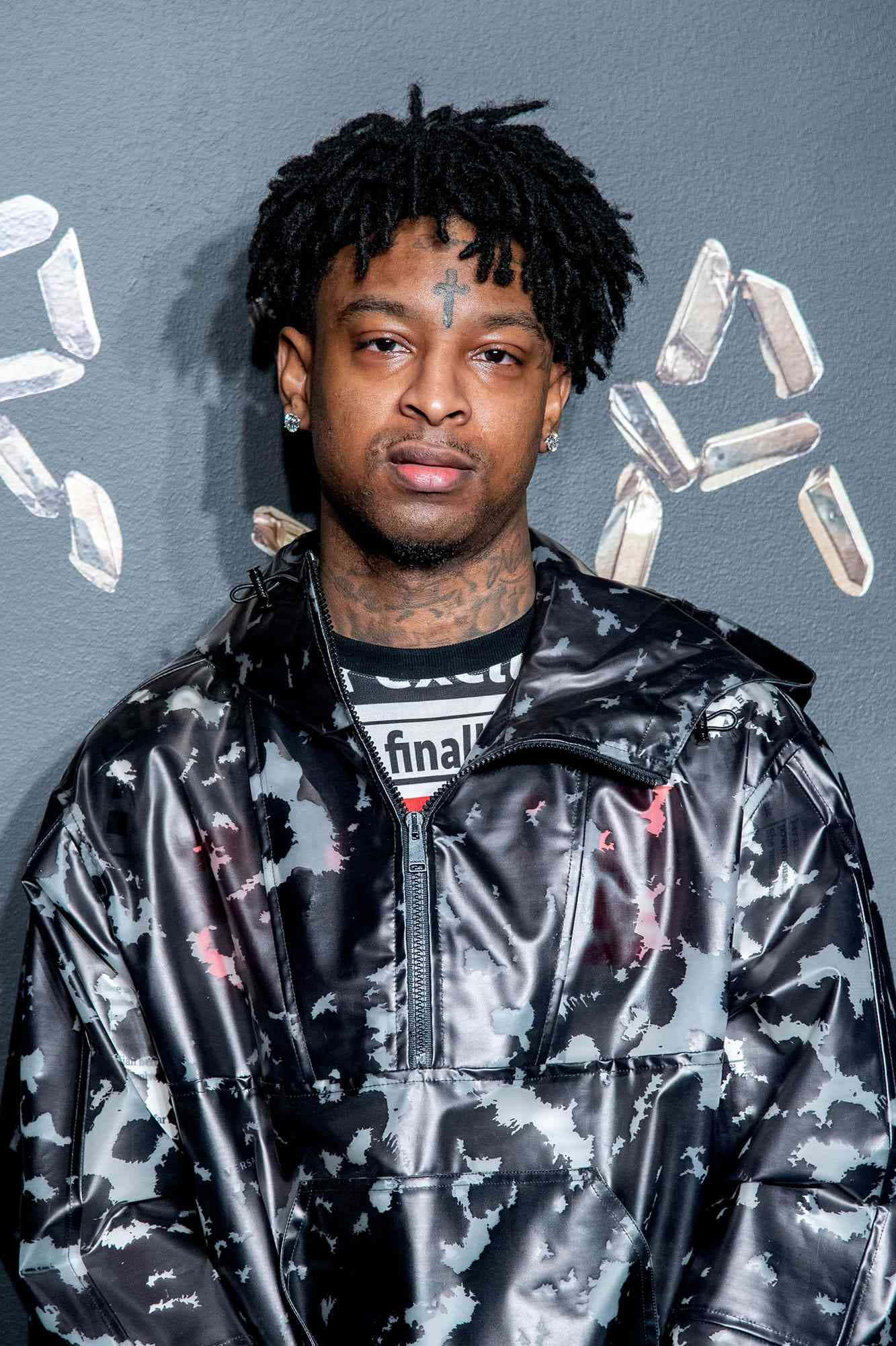 21 Savage 舞台演出 壁紙 — 一名留著辮子頭、帶有十字架紋身的男子，身穿閃亮迷彩外套，凝視前方，背景是帶有水晶貼紙的灰色牆壁。