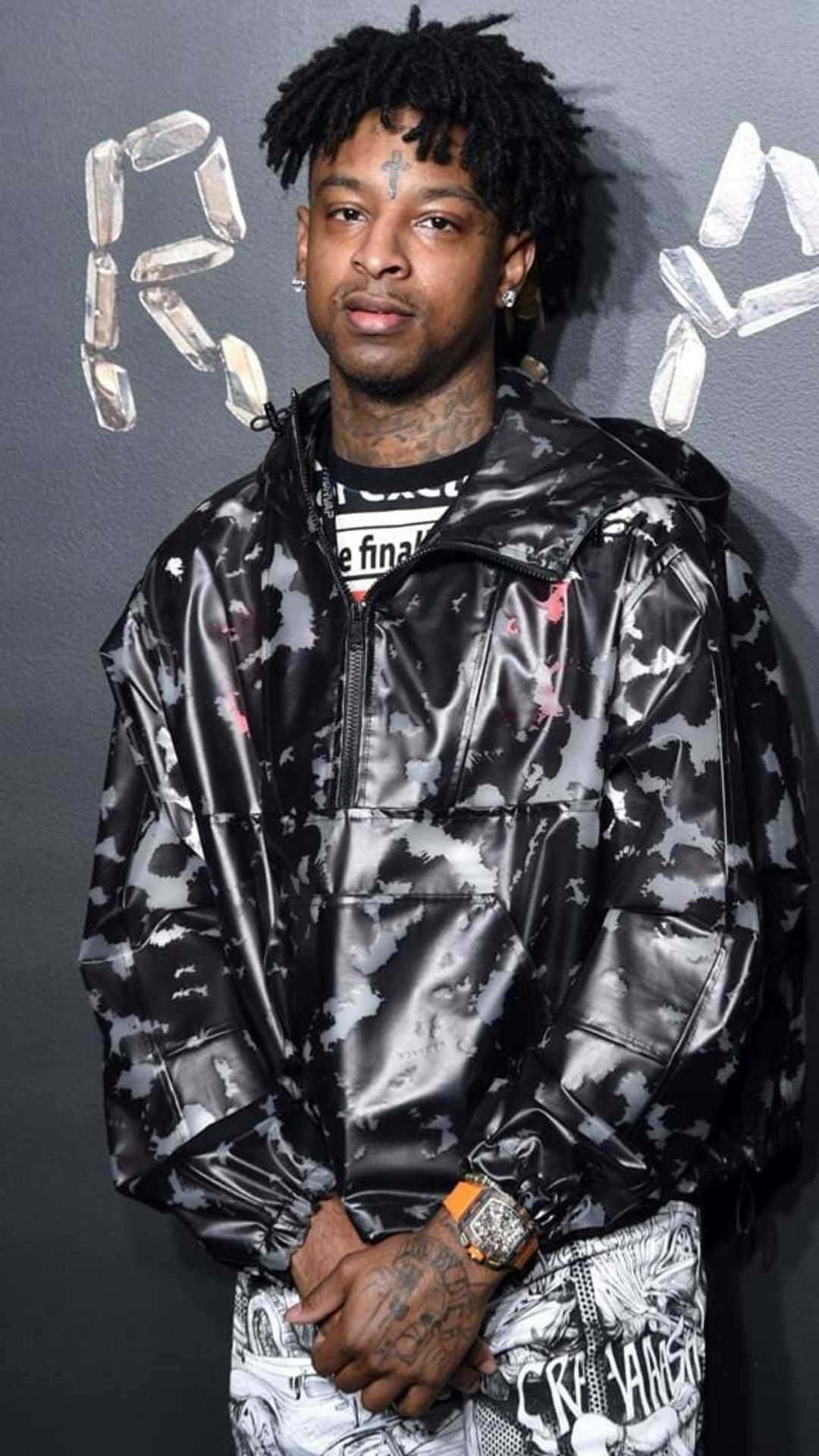 21 Savage 舞台演出 壁紙 — 一名留著辮子頭和紋身的男子，穿著閃亮的黑白迷彩外套，背景是灰色牆壁，上面有水晶般的字母。