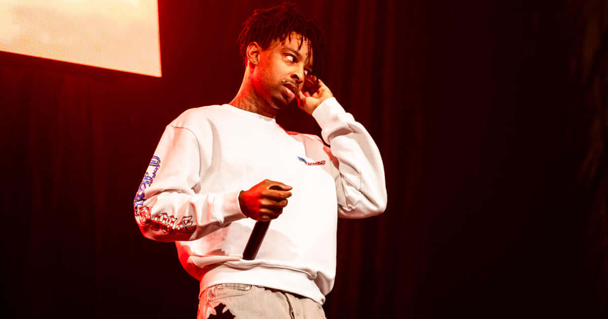 21 Savage 舞台演出肖像 壁紙 — 一位身穿白色運動衫的男性說唱歌手在溫暖的紅色舞台燈光下，以深色窗簾背景為襯，手持麥克風。