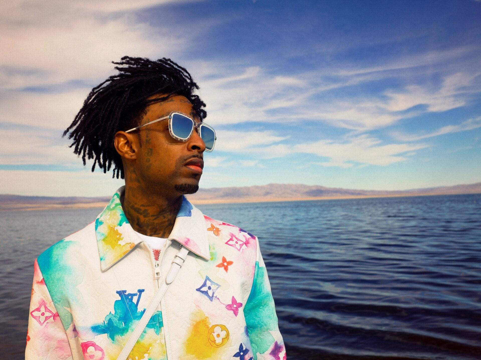 21 Savage 色彩繽紛 LV 夾克 壁紙 — 一名留著辮子頭、戴著太陽眼鏡的男子站在廣闊的藍色湖邊，天空局部多雲。