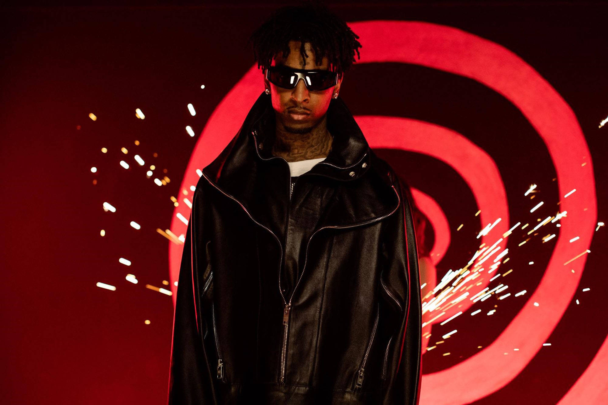 21 Savage 螺旋音樂錄影帶靜態畫面 壁紙 — 一名身穿黑色皮夾克和太陽眼鏡的男子站在紅色螺旋背景前，火花飛舞。