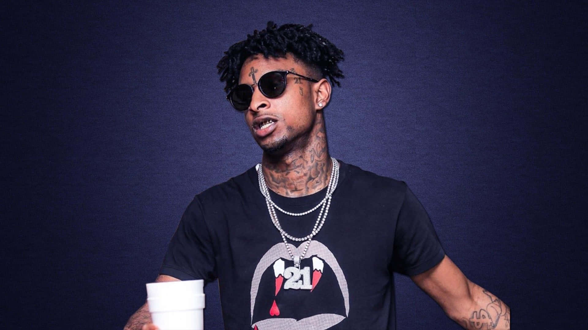 21 Savage 身穿標誌性紅黑裝束展現自信 壁紙 — 一個有紋身和太陽眼鏡的男人在深藍色工作室背景前拿著一個白色杯子。