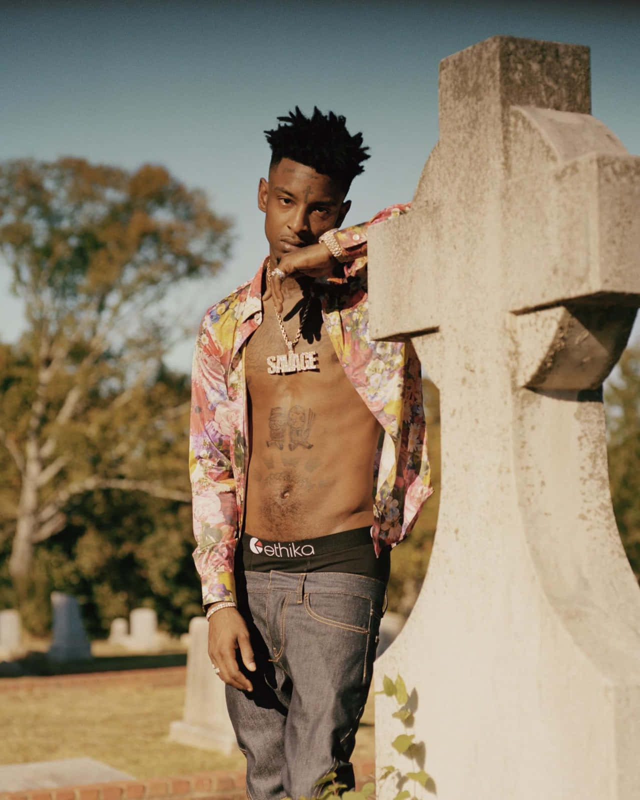 21 Savage | 音樂傳奇與熱門歌曲製造者 壁紙 — 一名身穿花卉圖案襯衫的男子，在陽光明媚的墓園裡靠著一塊風化的墓碑，背景有樹木。