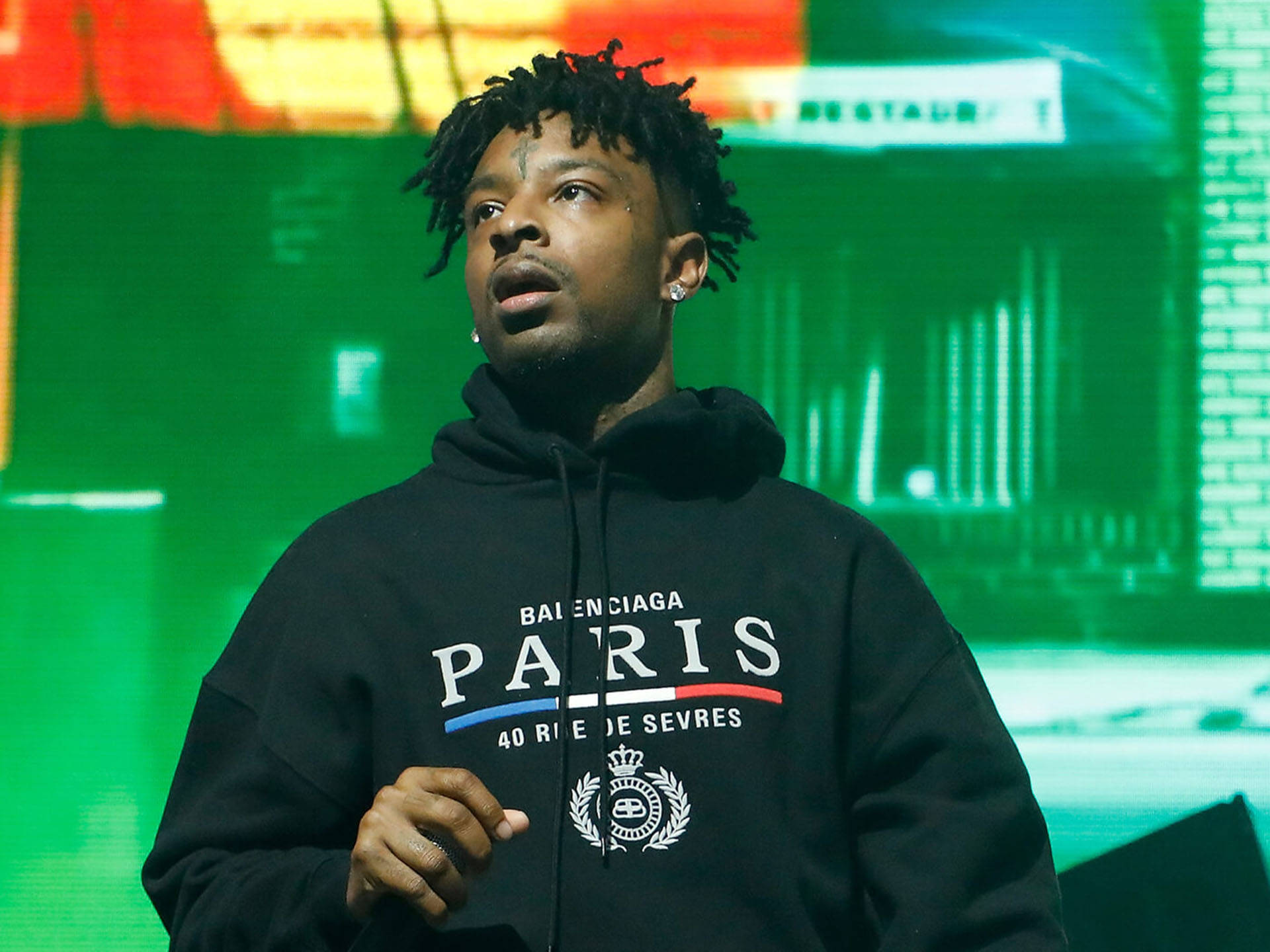 21 Savage出席Forbes 30 Under 30活動 壁紙 — 一名留著辮子、身穿黑色 Balenciaga Paris 連帽衫的男子站在舞台上，舞台被鮮豔的綠色和紅色燈光照亮。