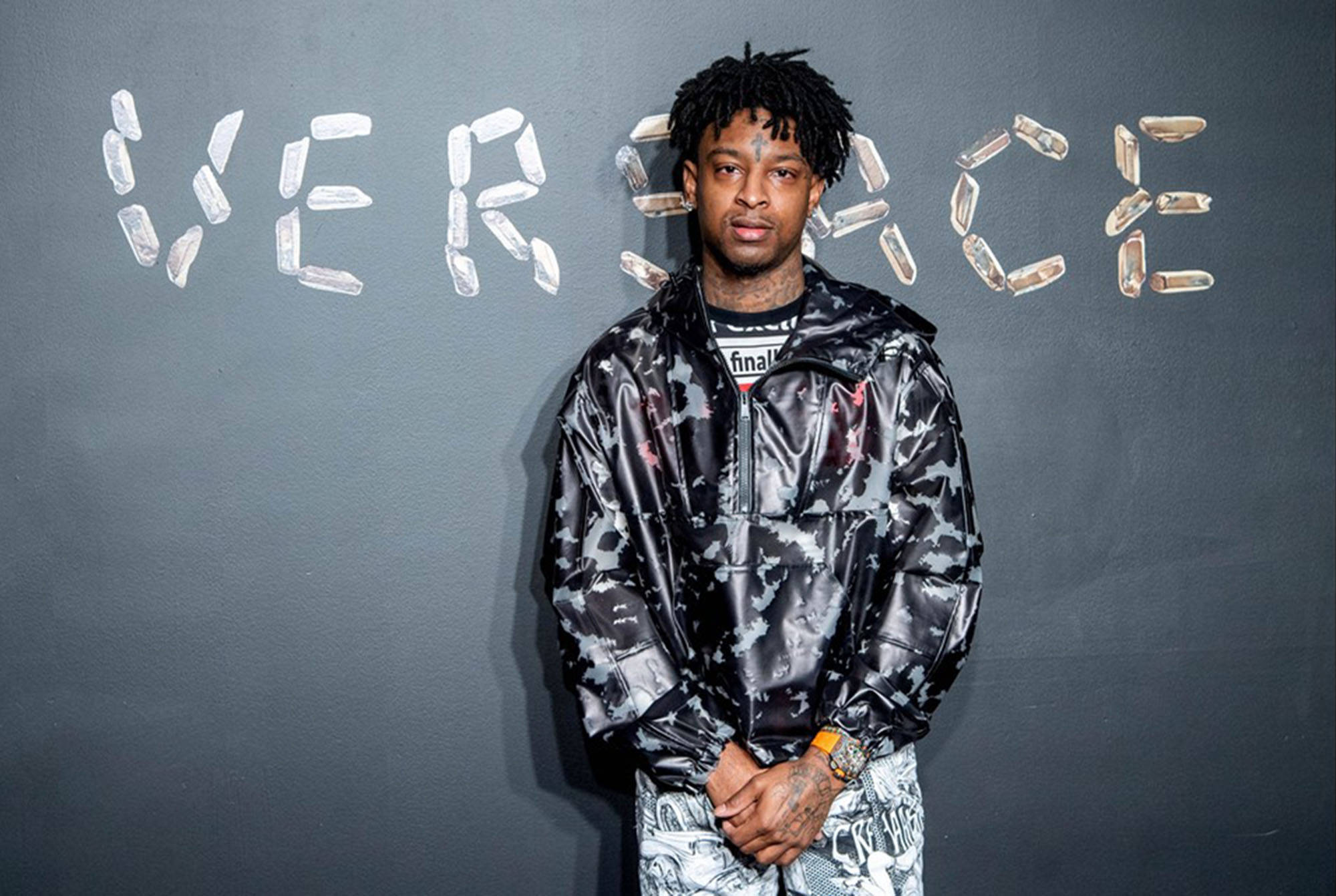 21 Savage出席Versace 2019秋冬秀 壁紙 — 一名留著辮子的男人自信地站在一件閃亮的迷彩外套中，背景是深色牆壁，上面用瓶蓋拼出「VERSACE」。