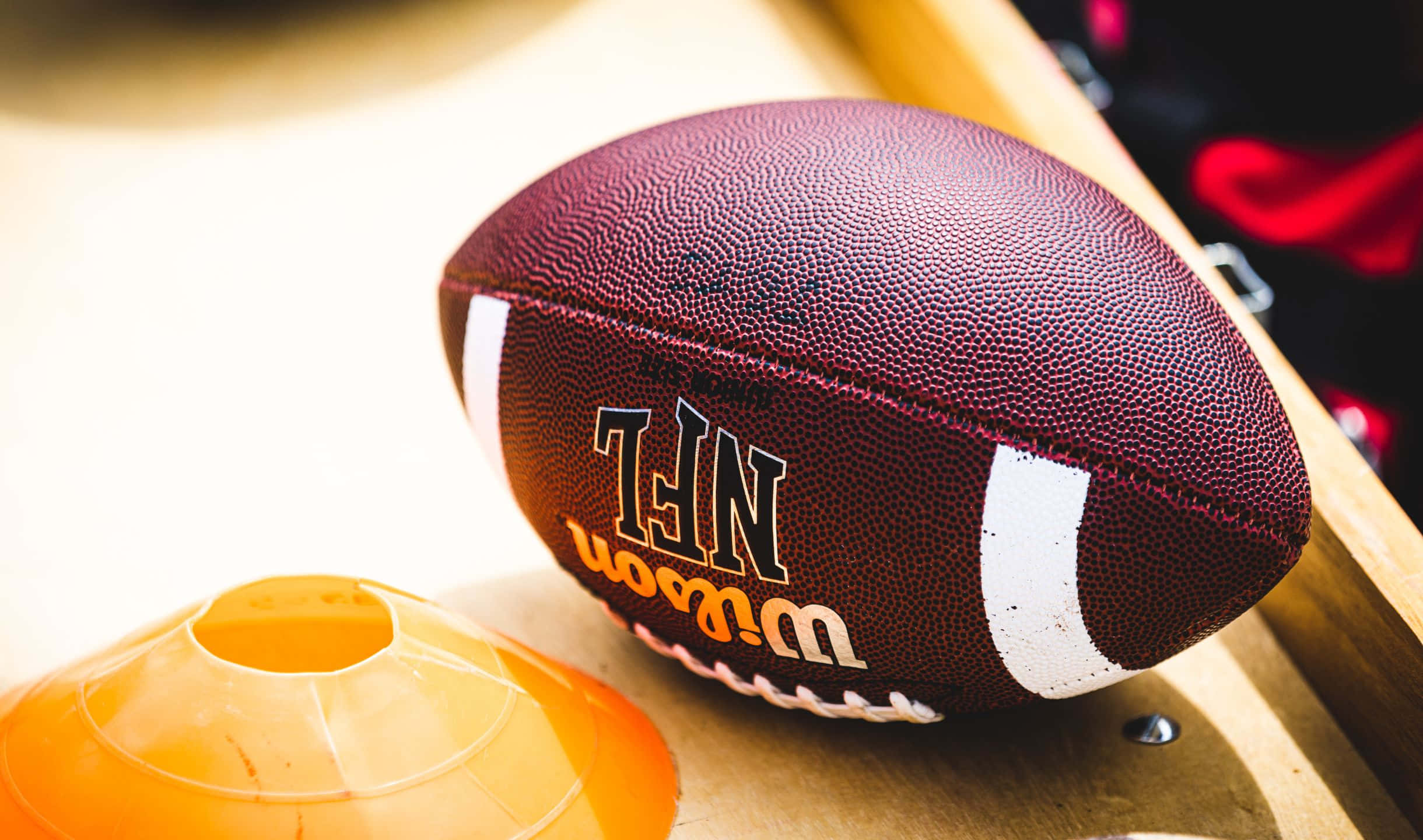 2440x1440 足球背景：Wilson NFL 橄欖球 壁紙 — 一個栗色NFL足球的特寫，旁邊放著一個橙色錐體在木質表面上。