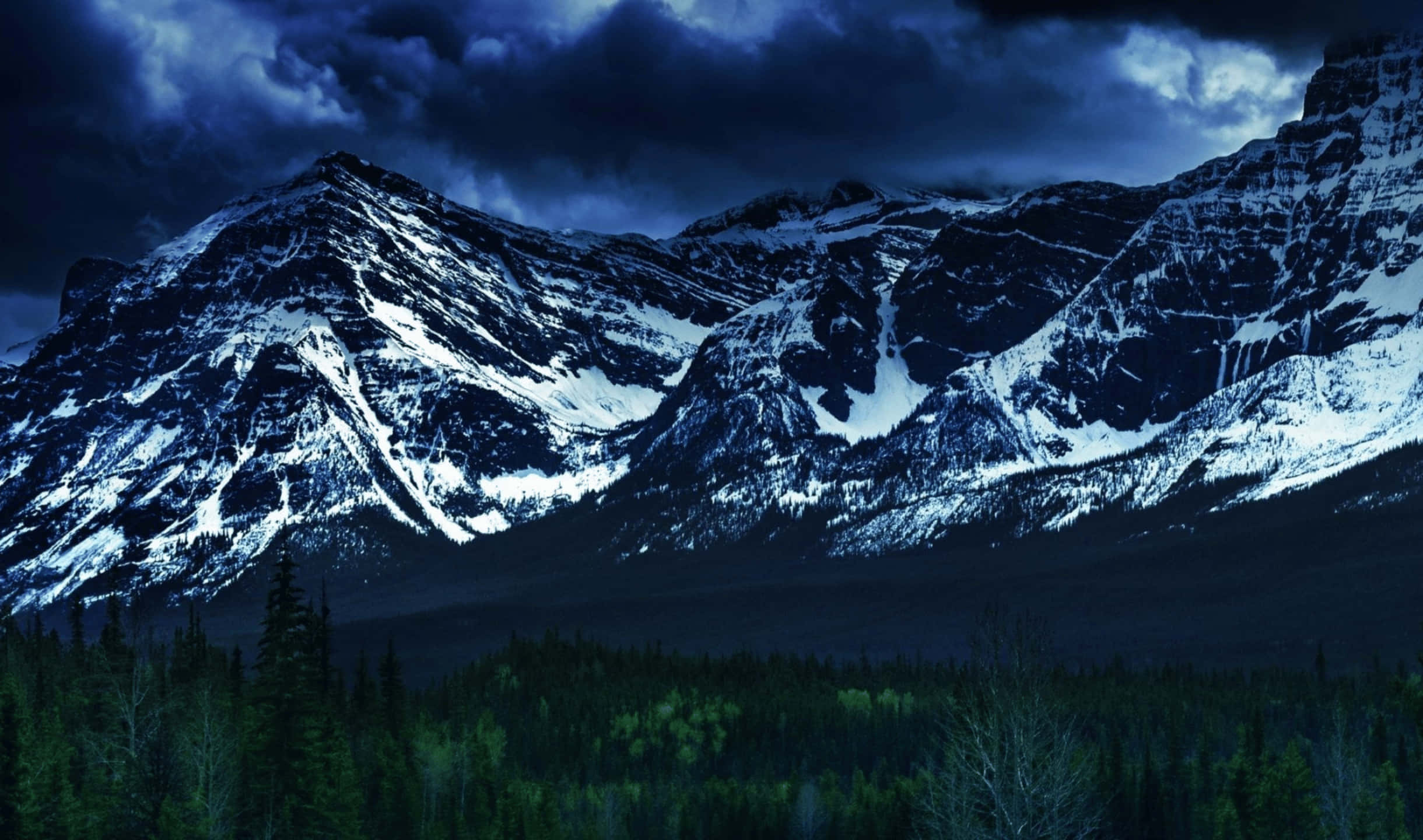 2440x1440 加拿大落基山監視器背景 壁紙 — 白雪覆蓋的山峰在風暴暮色天空下，聳立於茂密的常綠森林之上。