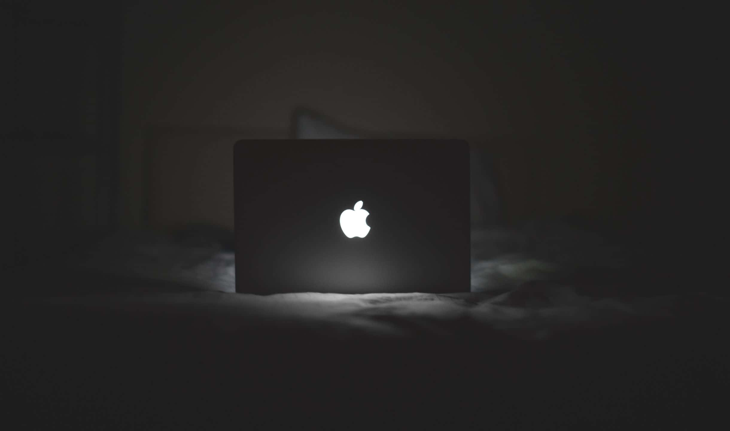 2440x1440 燈光 Apple 標誌背景 壁紙 — 一個發光的Apple標誌在關閉的筆記型電腦上，獨自放在昏暗房間的深色床上。