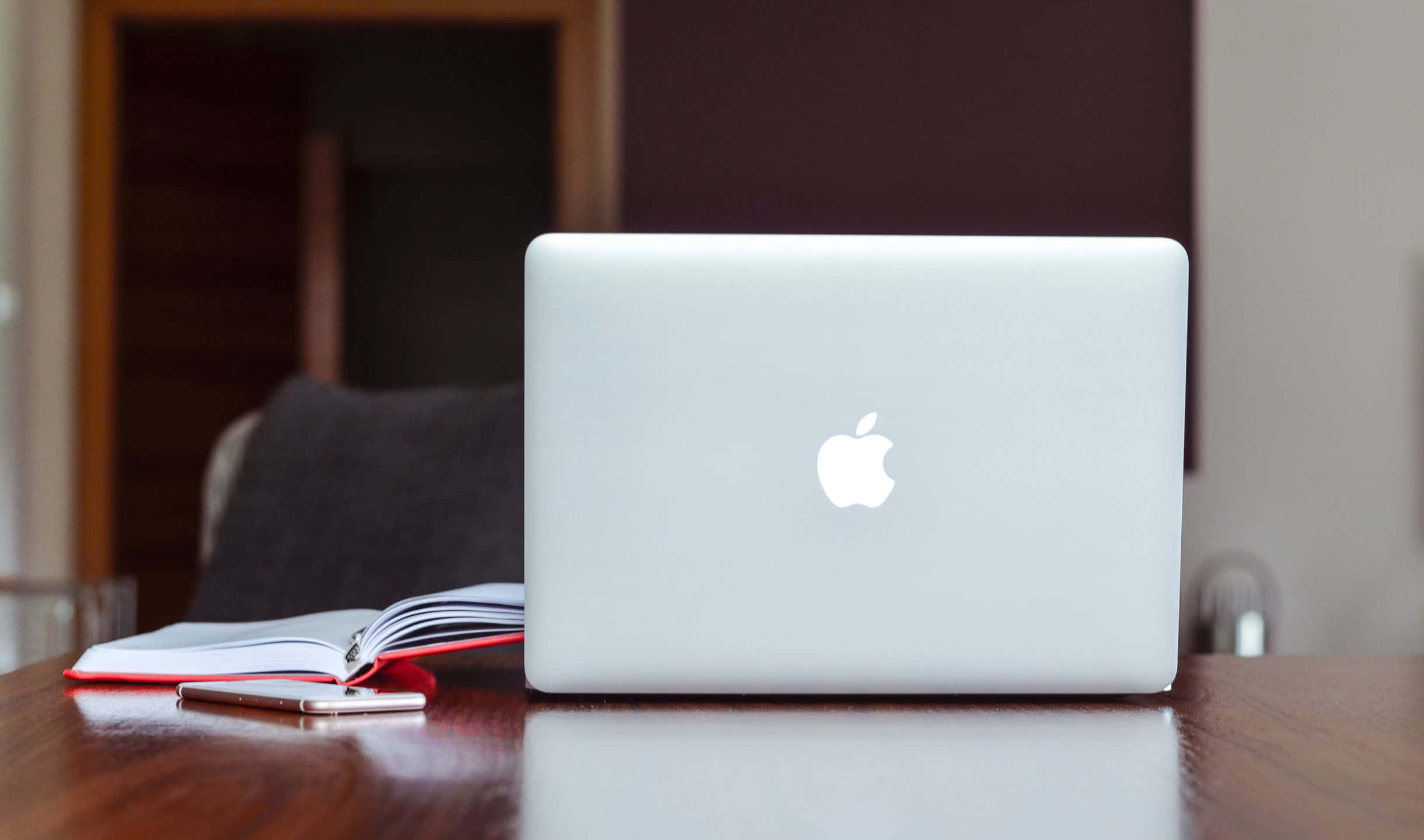 2440x1440 蘋果 MacBook 背景 壁紙 — 一部銀色手提電腦合上放在木桌上，旁邊有一本打開的筆記本和智能手機，房間光線柔和。
