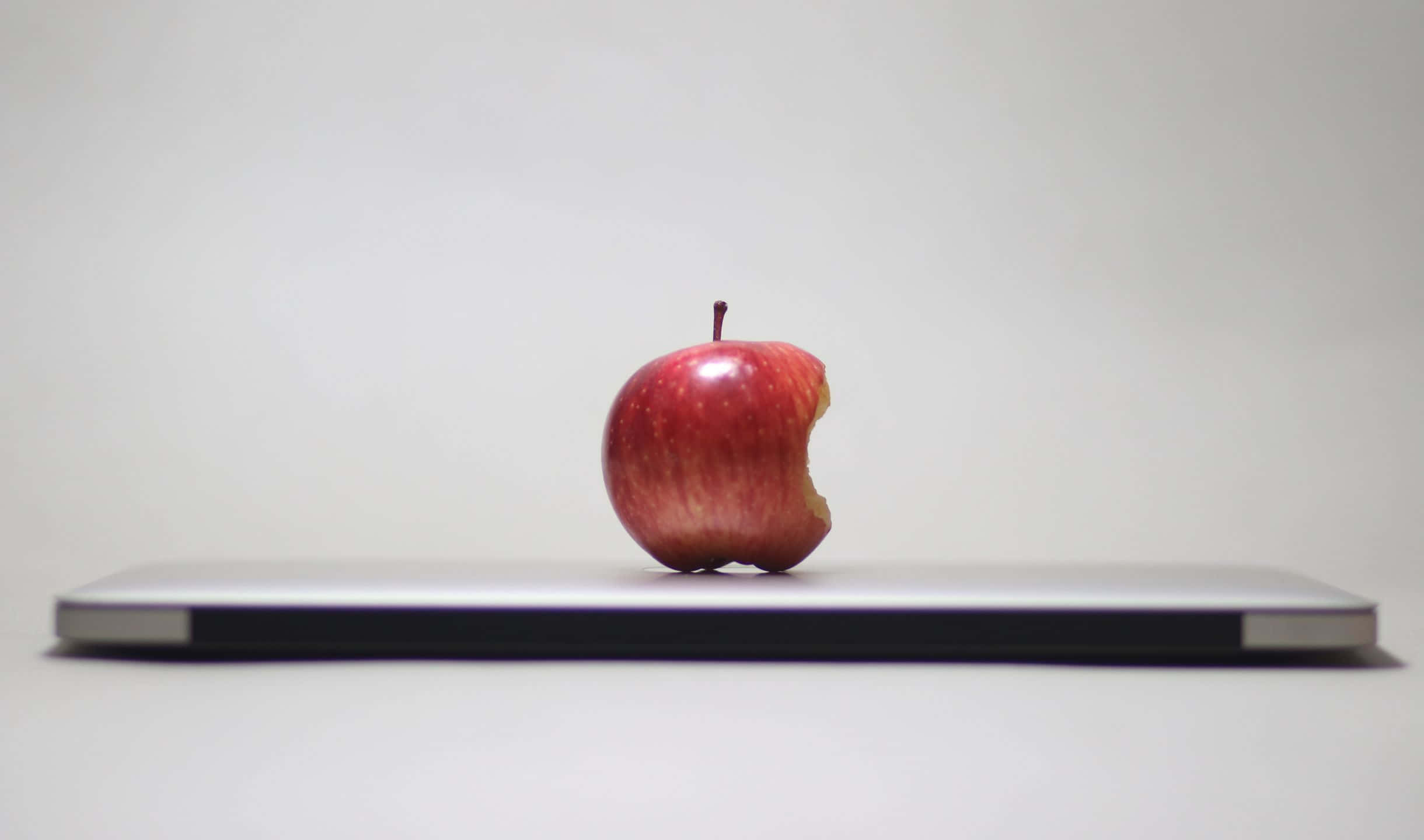 2440x1440 蘋果水果置於 MacBook 背景 壁紙 — 一個被咬過的紅蘋果放在關閉的銀色筆記本電腦中央，背景是純白色。