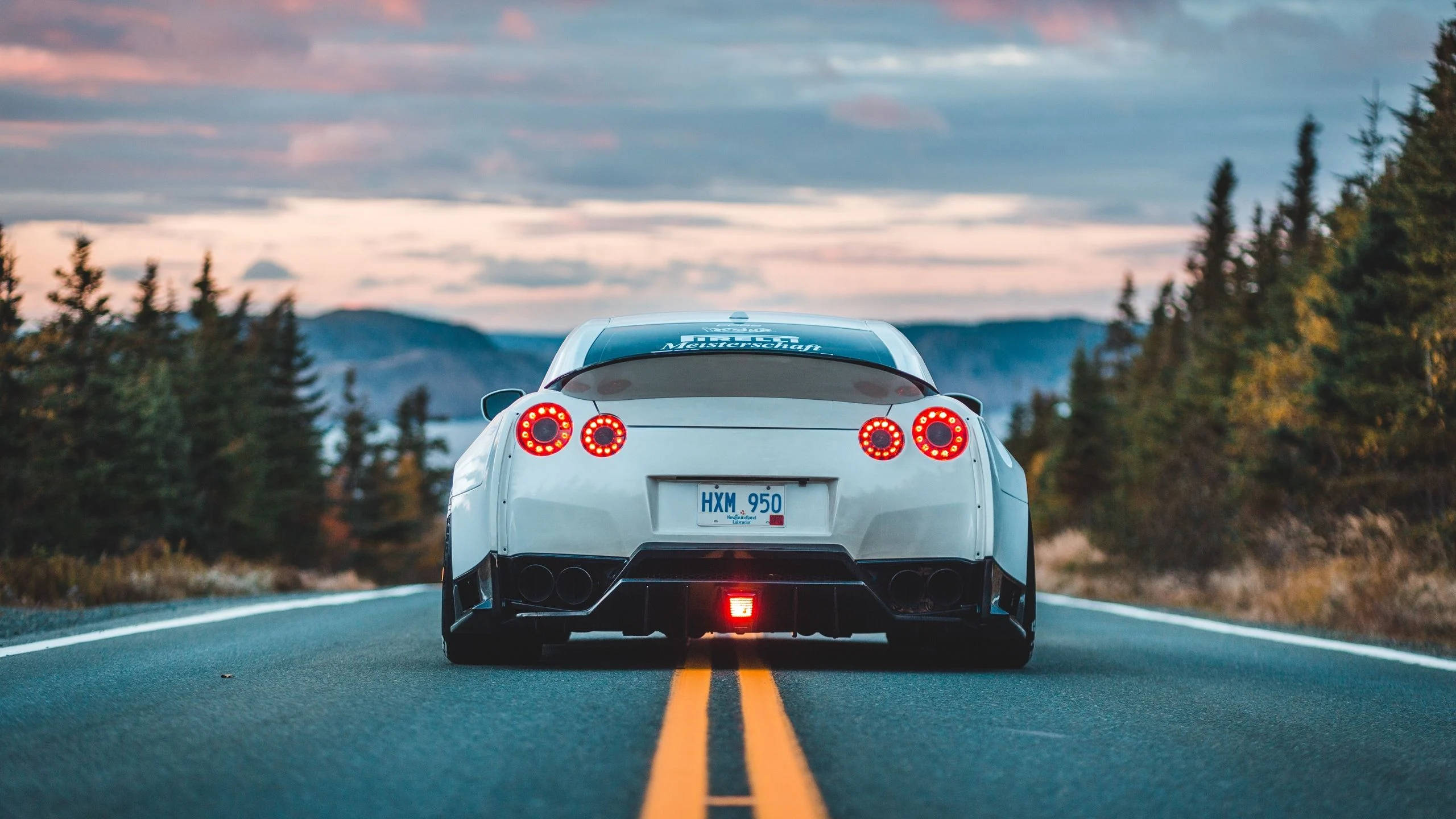 2560 X 1440 車款：白色 Nissan GT-R 壁紙 — 一輛白色跑車在黃昏時分行駛在森林環繞的道路上，紅色尾燈閃閃發光。