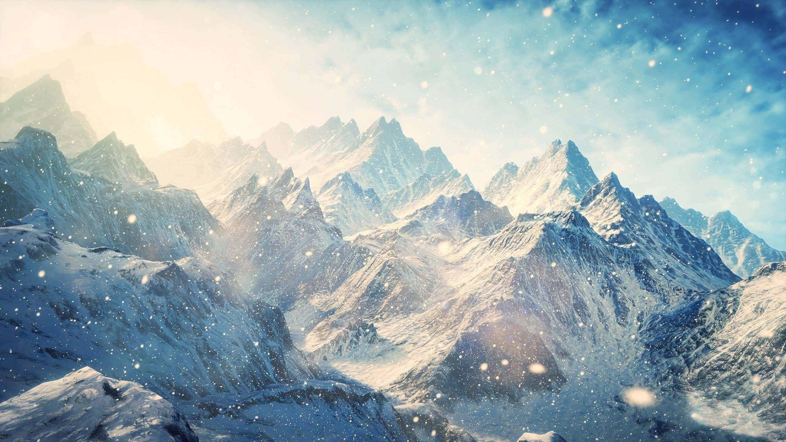 2560 X 1440 天際：冰封冬季山脈 壁紙 — 白雪覆蓋崎嶇的山峰，在柔和的藍天下，輕柔的雪花在空中飄舞。