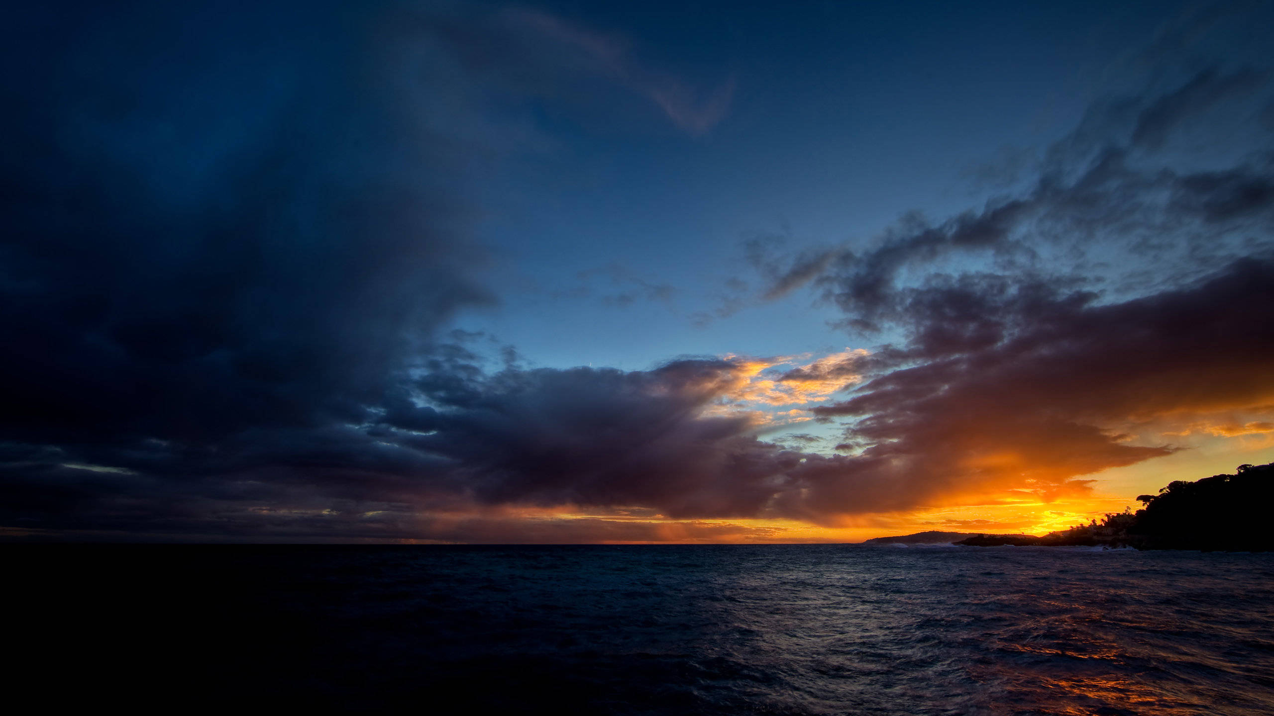2560x1440 風暴海景 壁紙 — 深藍色暮色天空下，黑暗海洋上空出現戲劇性的日落，伴隨著火紅色的橙色雲彩。