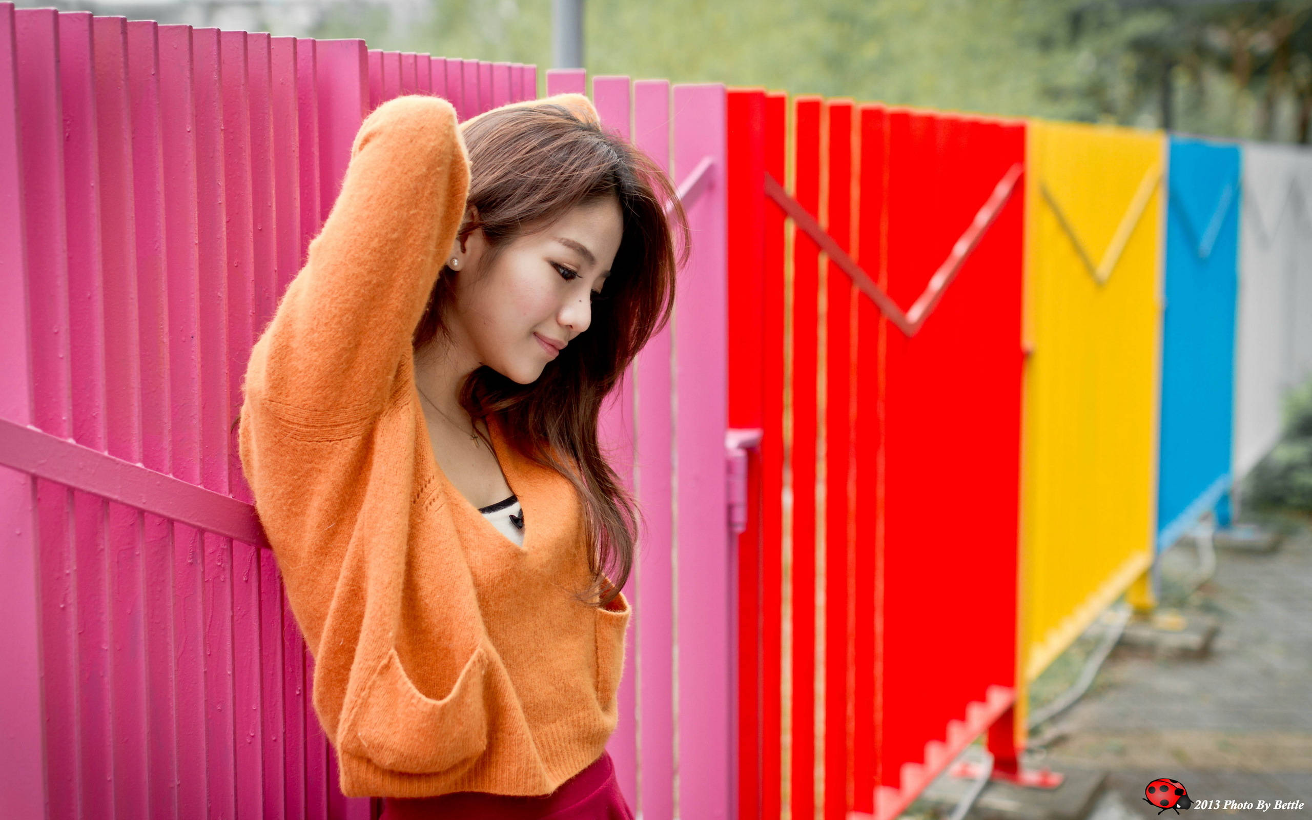 彩色柵欄旁女子 2560x1600 壁紙 — 一位穿著橙色毛衣的年輕女子，帶著溫柔的微笑，靠著充滿活力的彩虹色圍欄。