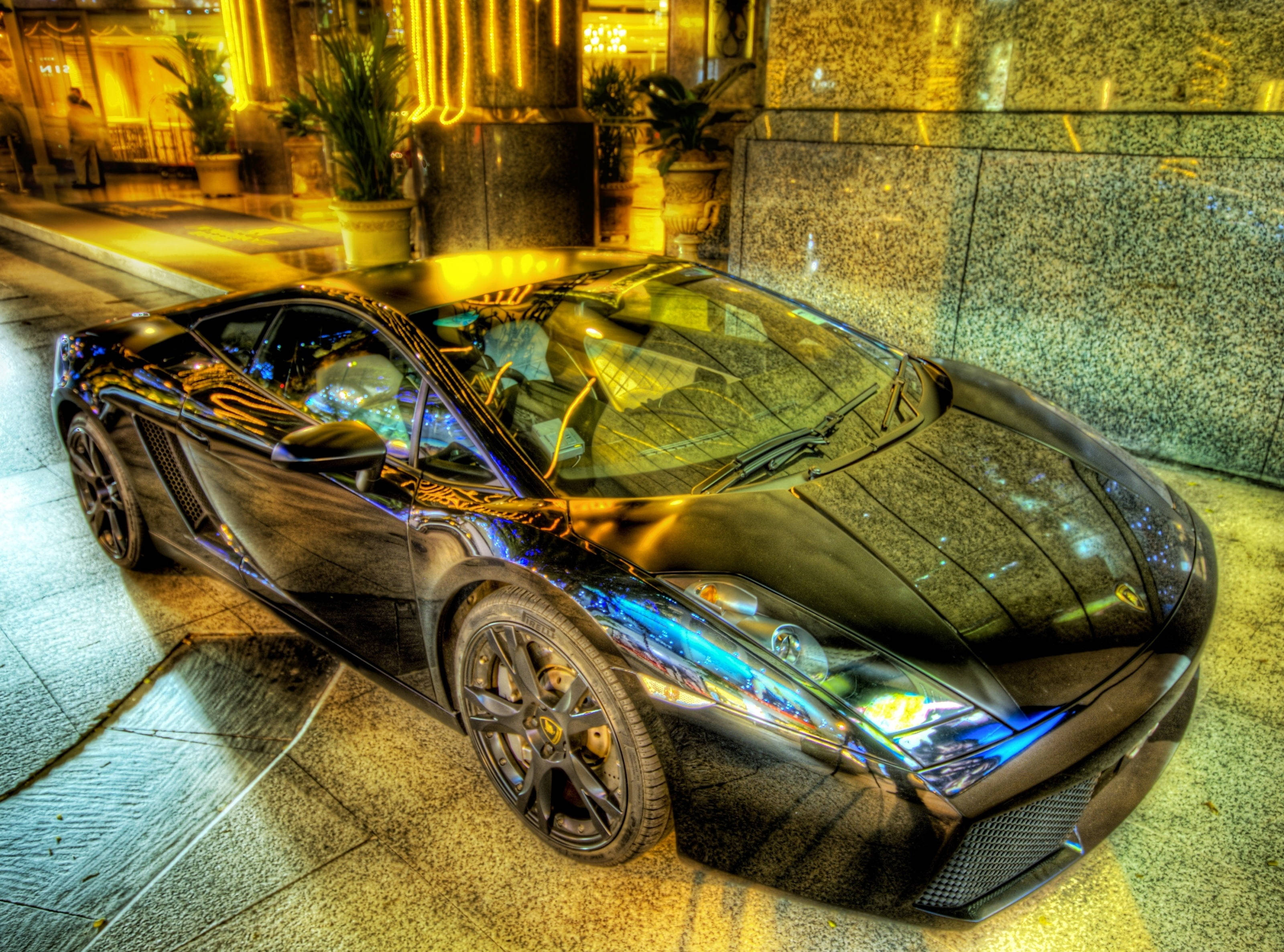2K 兰博基尼Aventador 壁紙 — 一輛光澤黑色跑車在夜間停泊在瓷磚城市人行道上，在溫暖的黃色街燈下閃閃發光。