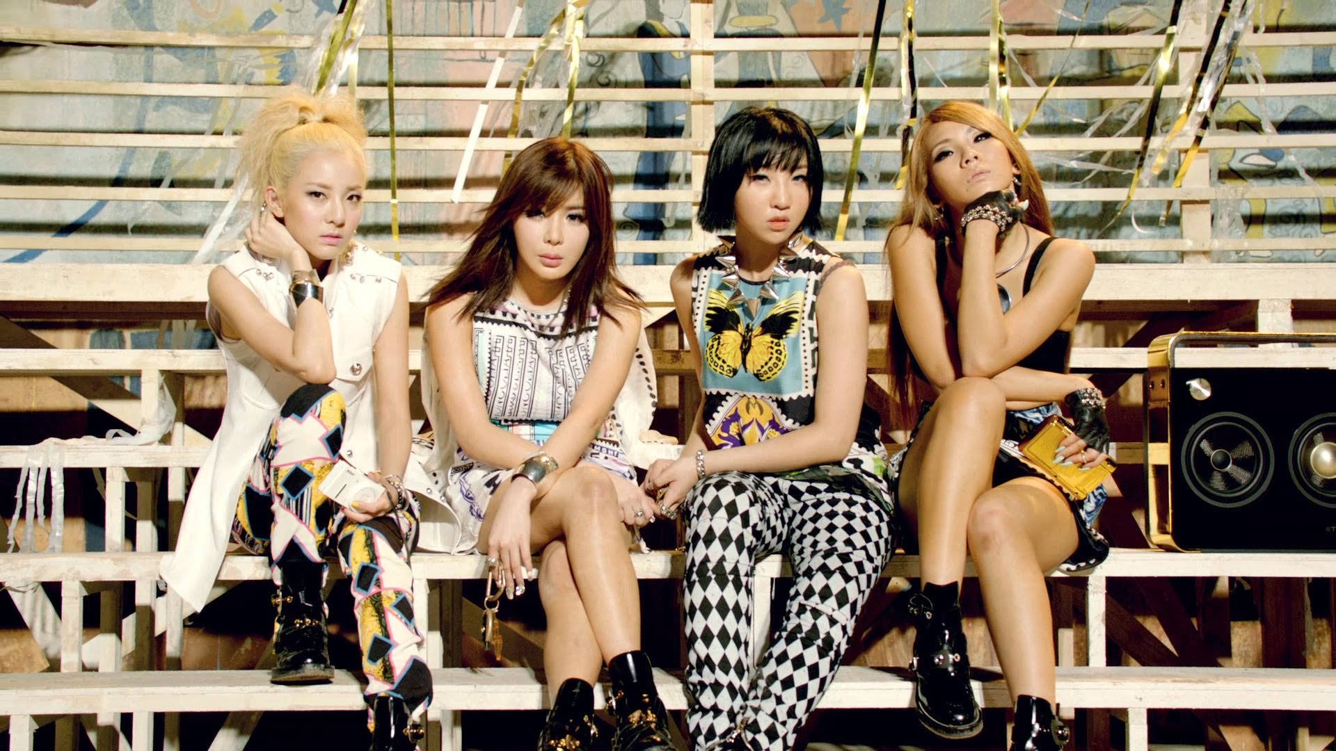 2NE1《Falling In Love》 壁紙 — 四位時尚女性穿著大膽圖案服裝，在風化的看台上自信擺姿勢，背景有塗鴉和揚聲器。