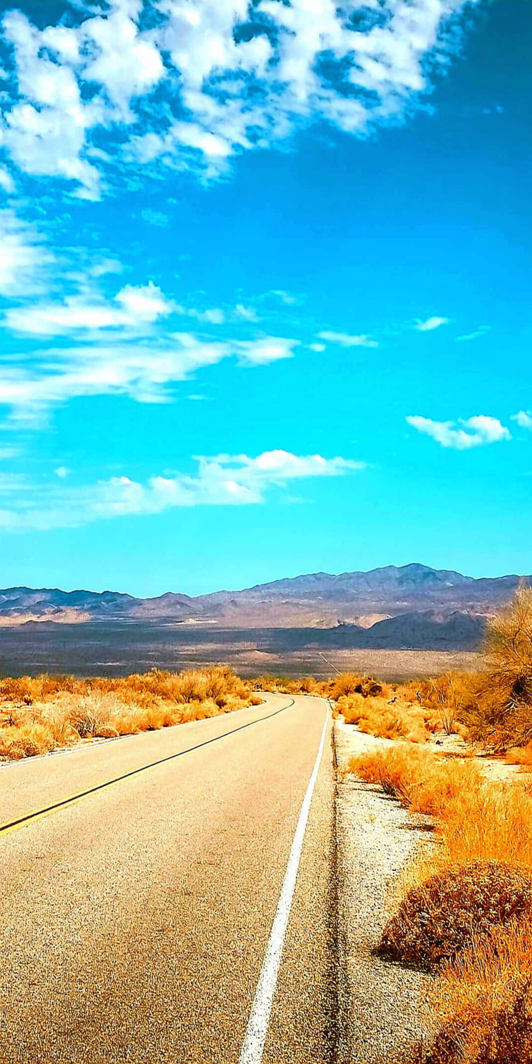像素3夏日背景：沙漠公路與棕草 壁紙 — 一條長直路穿過乾燥的沙漠景觀，上方是湛藍的天空，點綴著稀疏的白雲。
