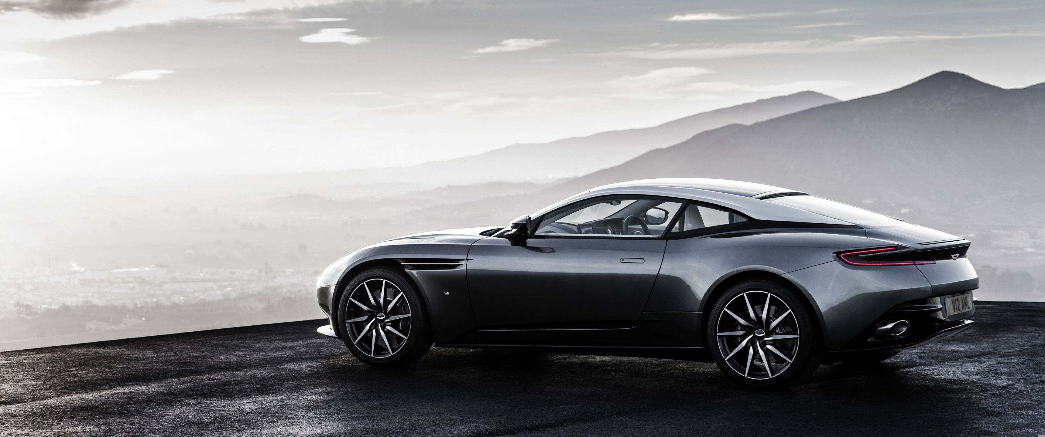3440x1440 賽車壁紙：阿斯頓·馬丁 DB11 壁紙 — 一架時尚的銀色跑車停在懸崖上，俯瞰著柔和天空下的朦朧山脈。