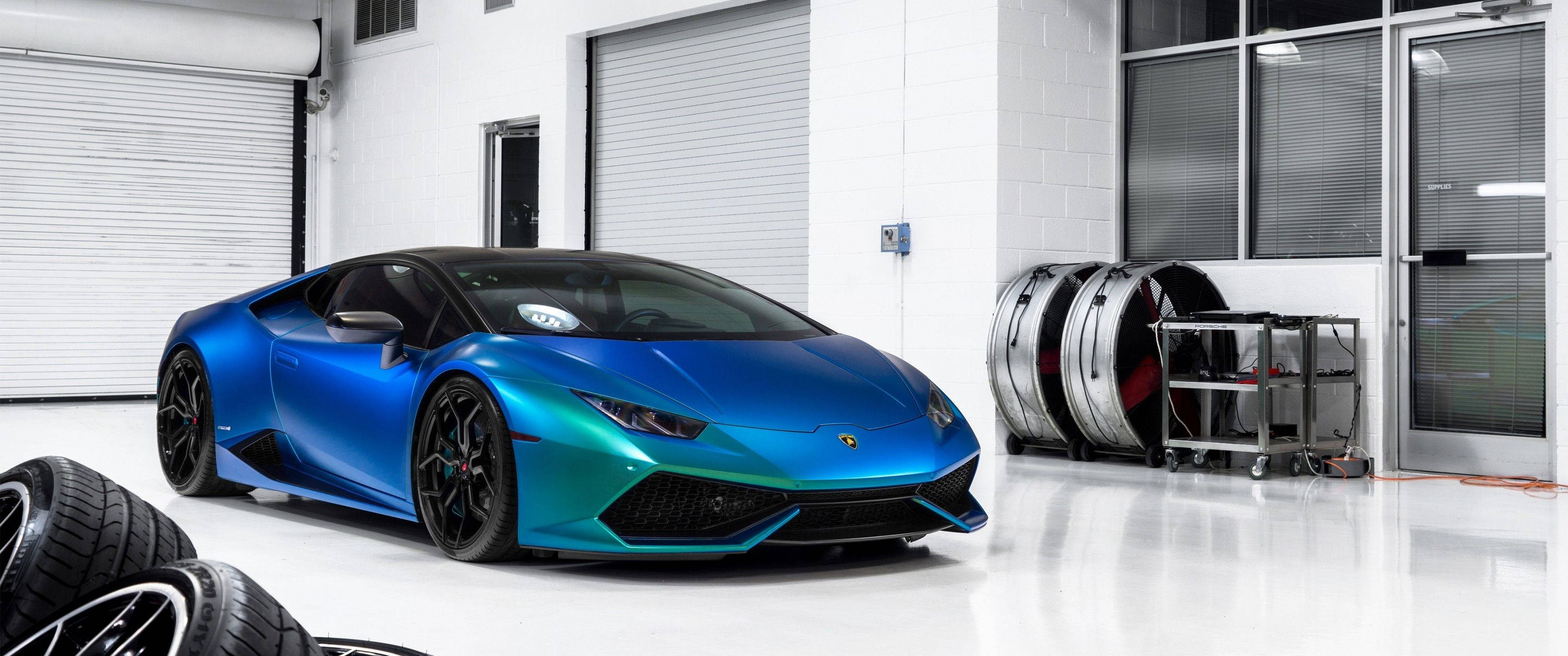 3440x1440 賽車壁紙：蘭博基尼 Huracan 壁紙 — 一輛充滿活力的藍色和藍綠色林寶堅尼Huracán跑車在明亮的燈光下閃耀，車庫潔白乾淨，堆疊著輪胎和工業設備。