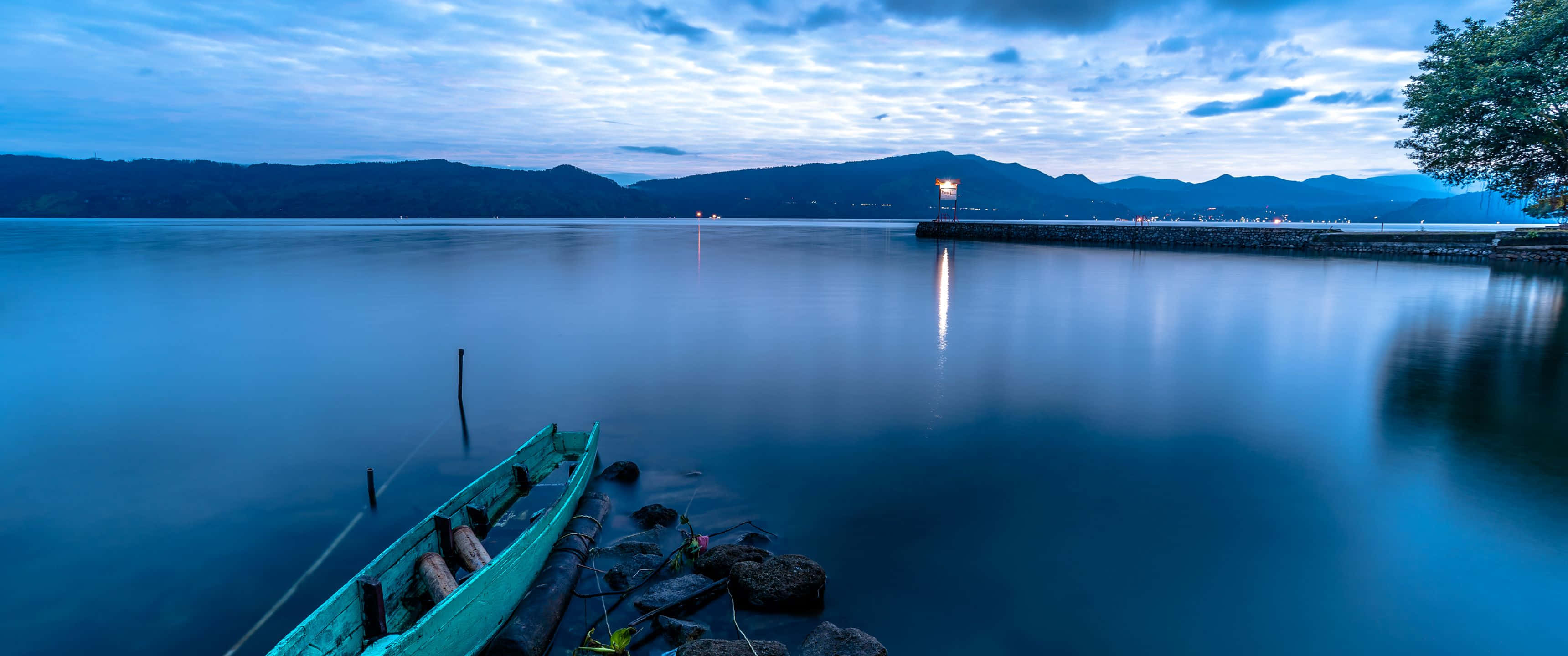令人著迷的 3440x1440 高畫質壁紙 壁紙 — 一艘青綠色小船在黃昏時停泊在岩石海岸上，平靜的藍色水面倒映著遠山和發光的碼頭燈光。