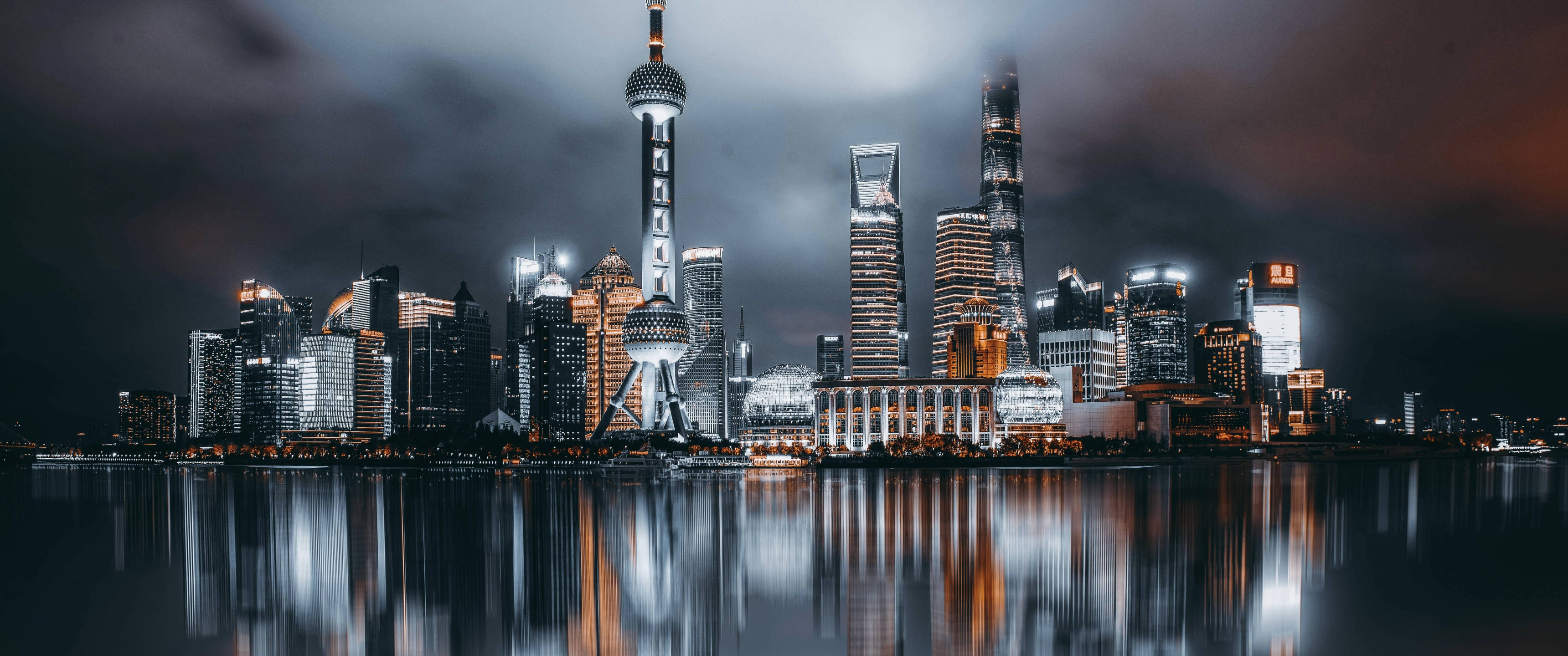 3440x1440 上海夜景 壁紙 — 上海天際線的戲劇性夜景，雲層密布的天空下，燈火通明的摩天大樓倒映在漆黑的水面上。