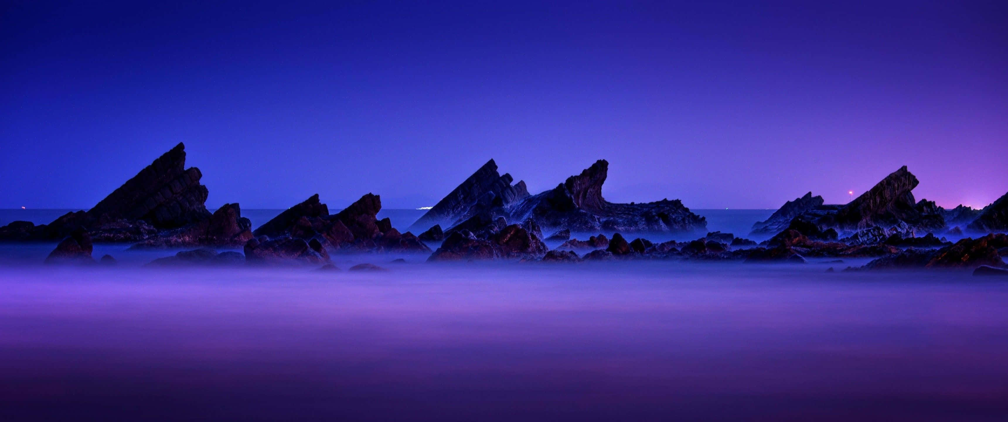最佳 3440x1440 高清風景 壁紙 — 在深沉的暮色天空下，輪廓分明的鋸齒狀岩石從霧氣瀰漫的紫色海洋中升起。