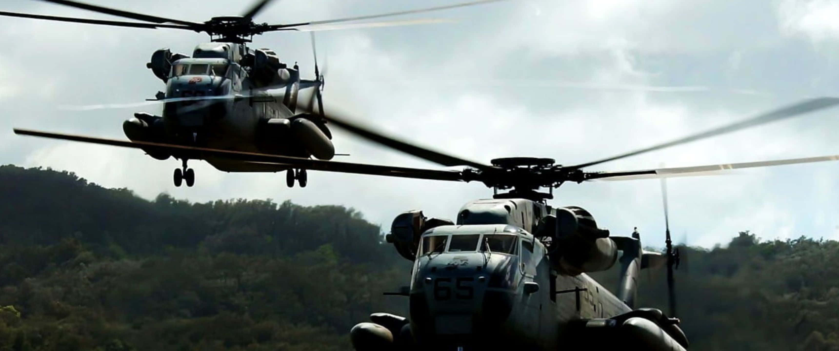 3440x1440p 直升機上的乘客享受絕美空中景觀 壁紙 — 兩架軍用直升機在陰天籠罩下盤旋於樹木繁茂的山坡上空。