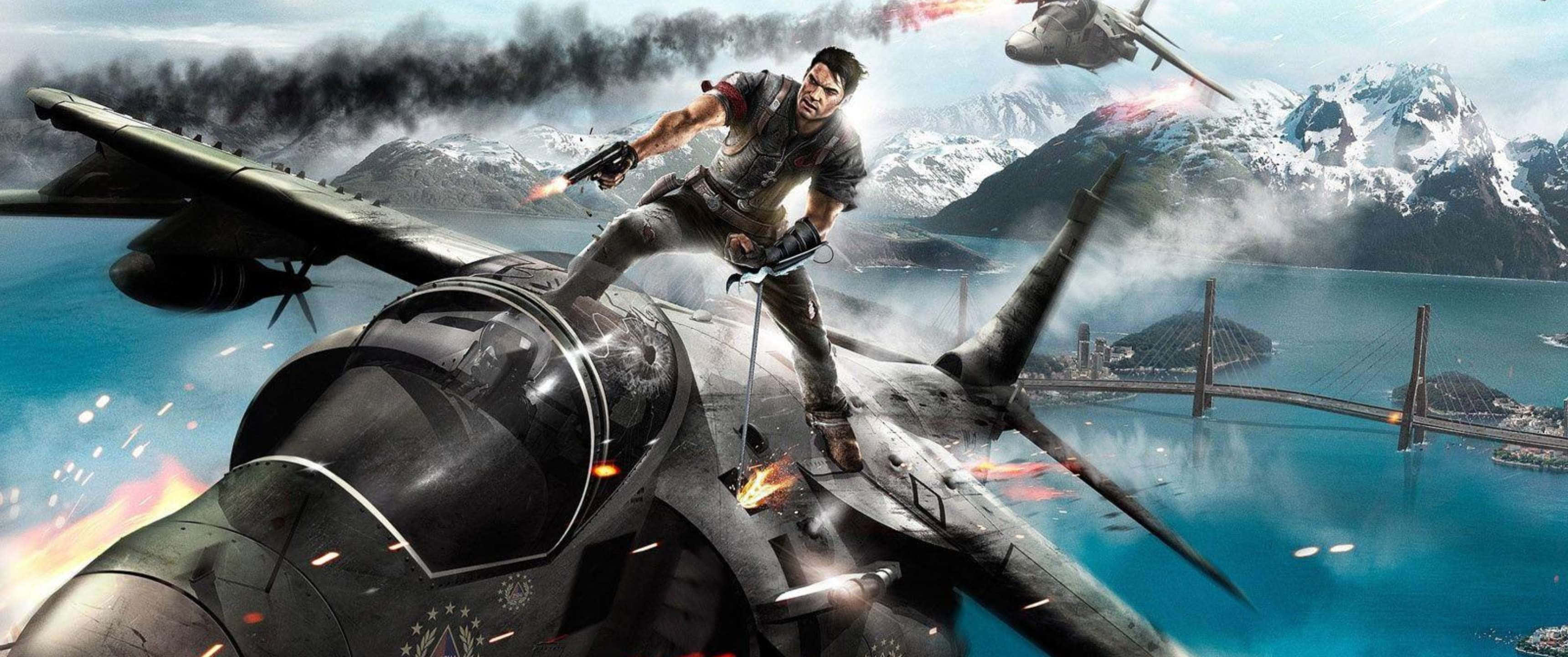 3440x1440p《Just Cause 4》主角背景 壁紙 — 一名士兵在爆炸中挑釁地站在戰鬥機上，下方是白雪覆蓋的山脈和一座橋樑。