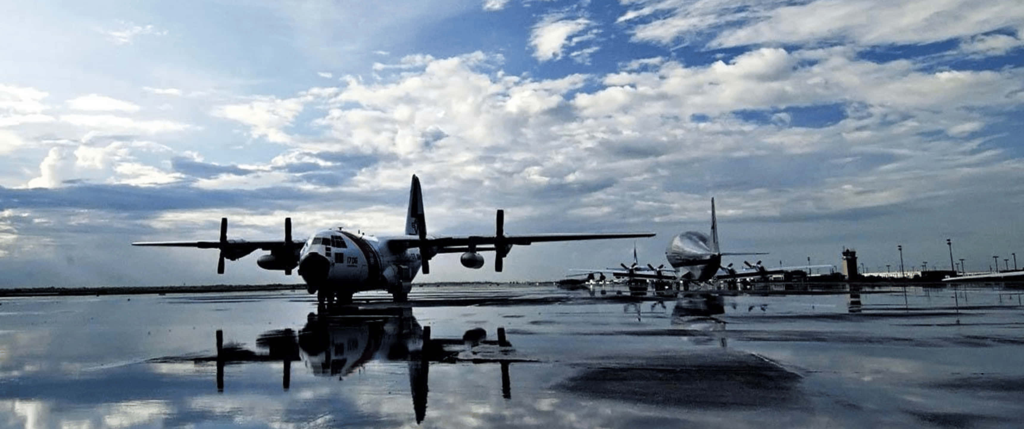 3440x1440p 停泊戰鬥機背景 壁紙 — 復古軍用飛機在陰雲密布的天空下形成剪影，倒映在機場濕漉漉的停機坪上。