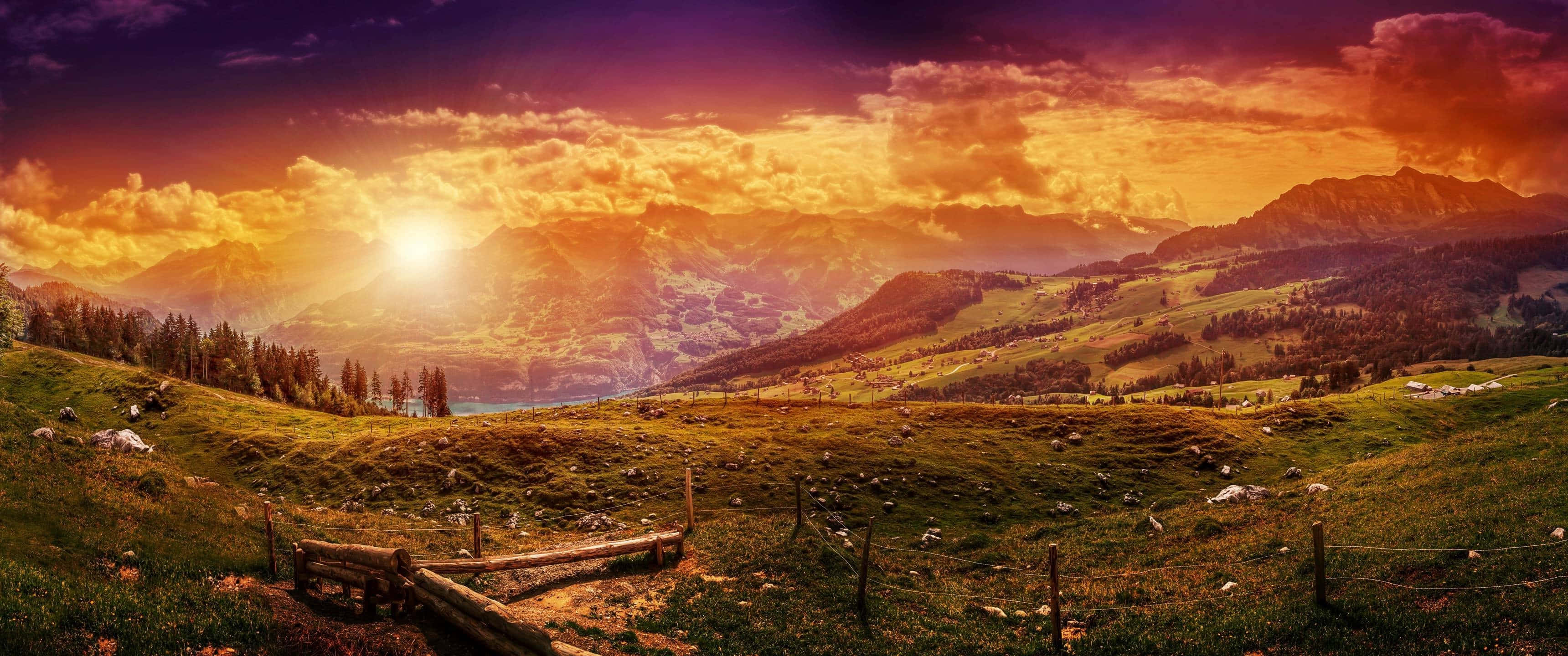3440x1440p 山脈與夕陽天空背景 壁紙 — 令人驚嘆的日落，映照著起伏的綠色山丘，遠方有山脈，前景是木柵欄。