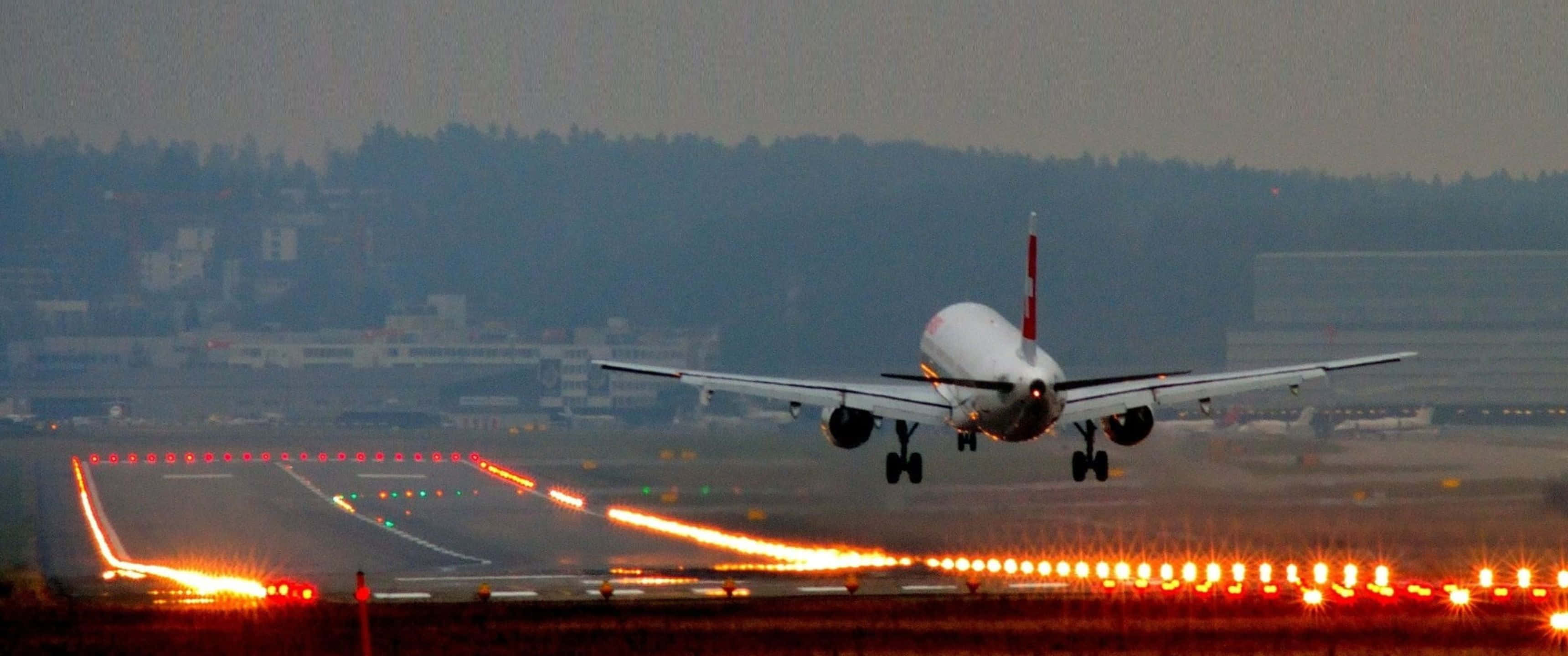 3440x1440p 巨型客機橙色燈光跑道背景 壁紙 — 一架大型飛機在黃昏時起飛，發光的跑道燈延伸至朦朧的遠方。