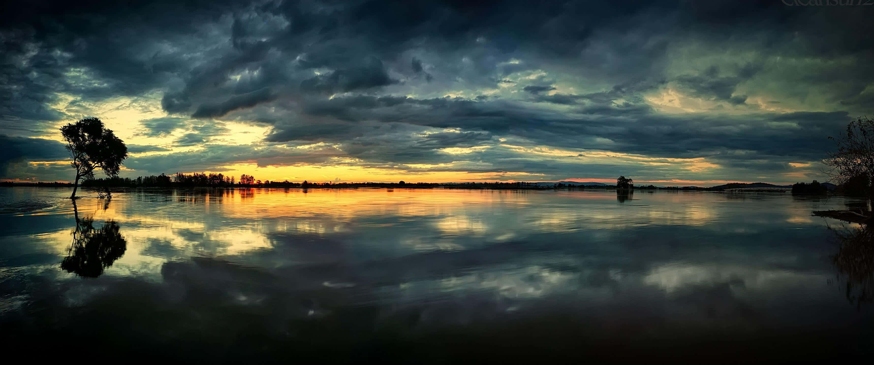 3440x1440p 暗雲天空背景 壁紙 — 一棵孤獨的樹矗立在平靜的水面上，映照出帶有烏雲和金色地平線的戲劇性日落天空。