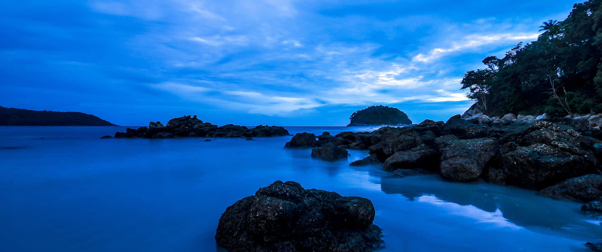 多雲天空下，黃昏時分平靜藍色海水中的深色岩石