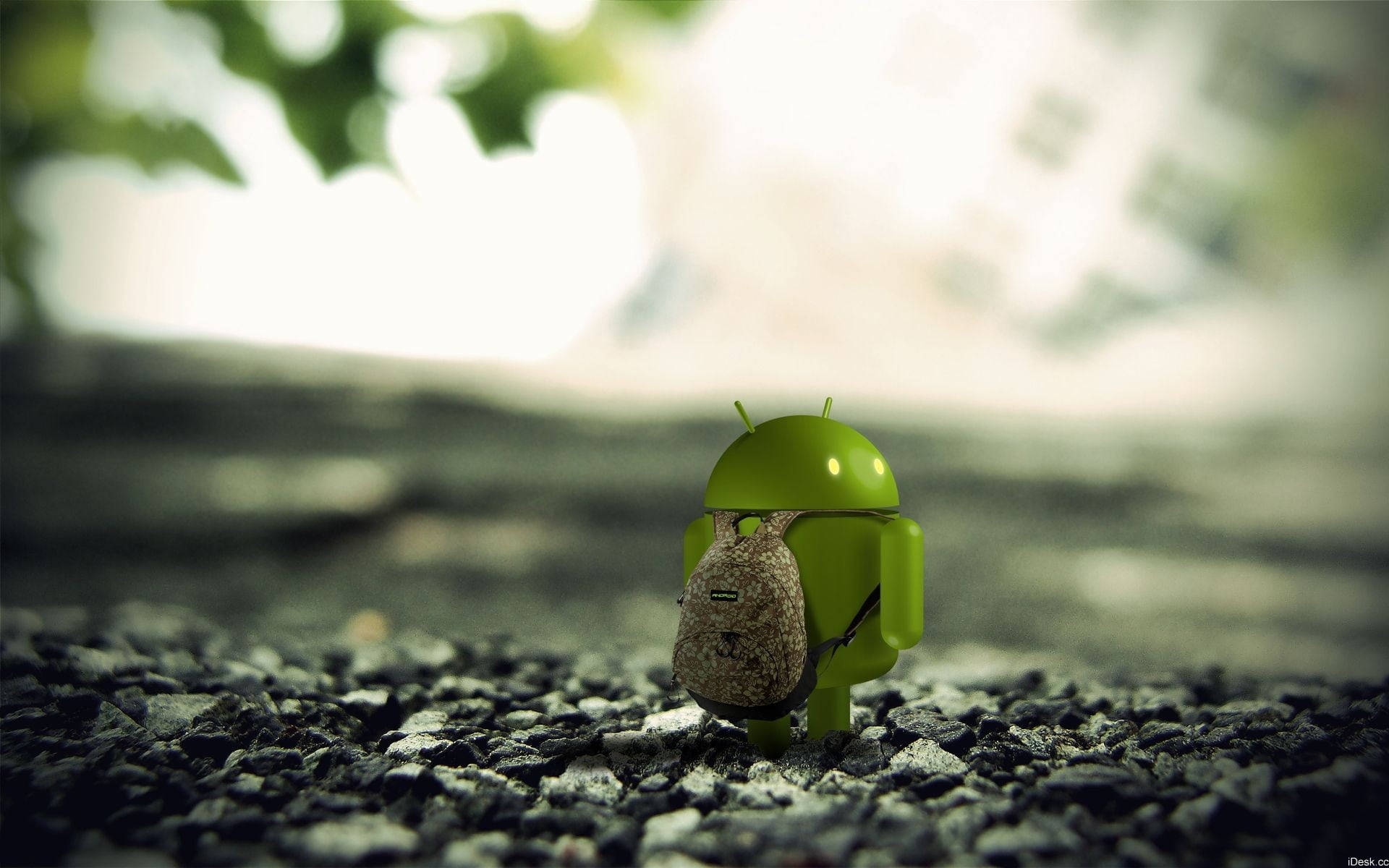 3D Android Logo 戴著 Android 背包的桌面壁紙 壁紙 — 一個細小的綠色Android公仔揹著背包，在柔和自然光下站在碎石上，背景有模糊的樹葉。