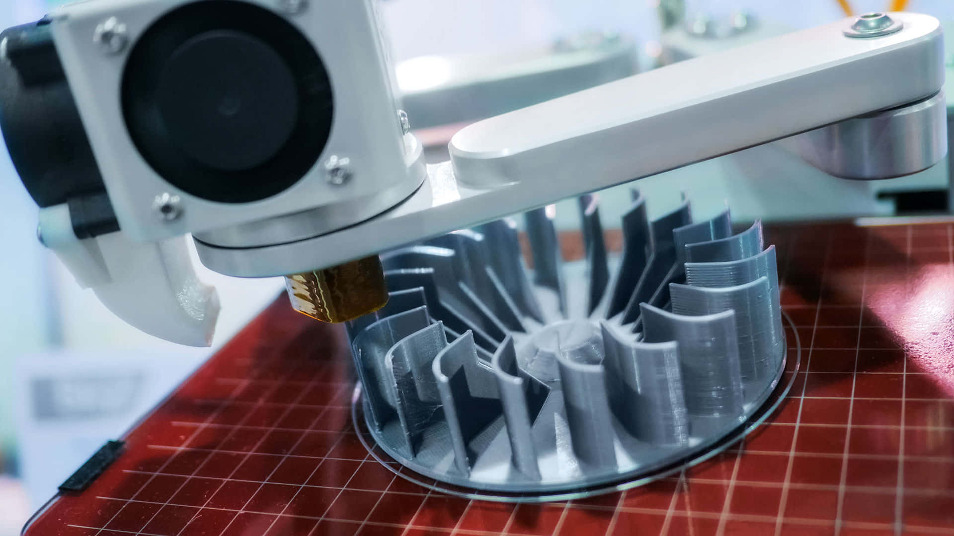 3d Printer 桌布 壁紙 — 3D打印機特寫，正在擠出灰色線材，在紅色網格床上構建一個風扇形狀的物體。