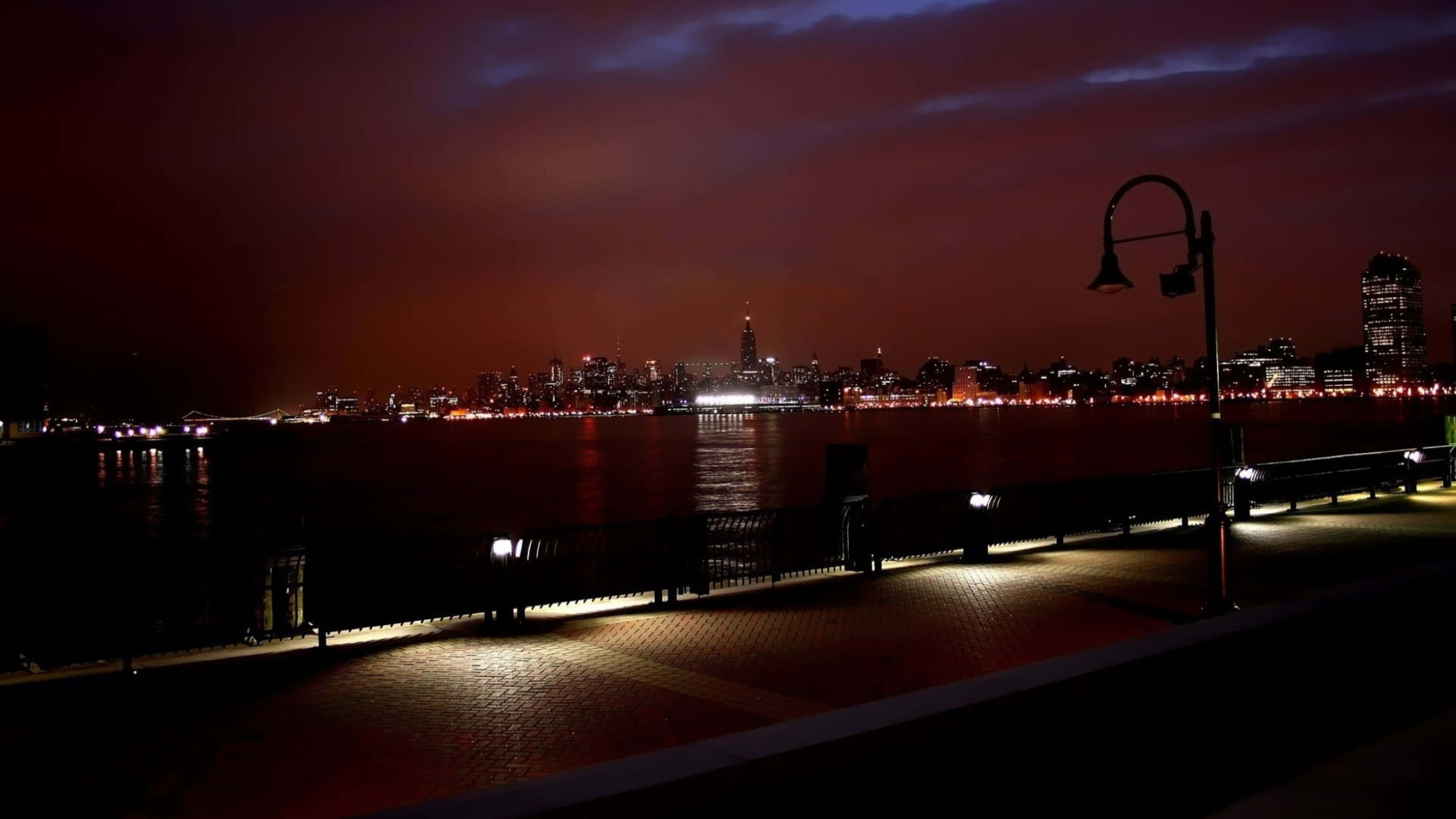 4K 紐約市灣畔公園夜景 壁紙 — 夜晚的海濱長廊，有發光的街燈和遠處的城市天際線，在深紅色的天空下倒映在黑暗的水面上。