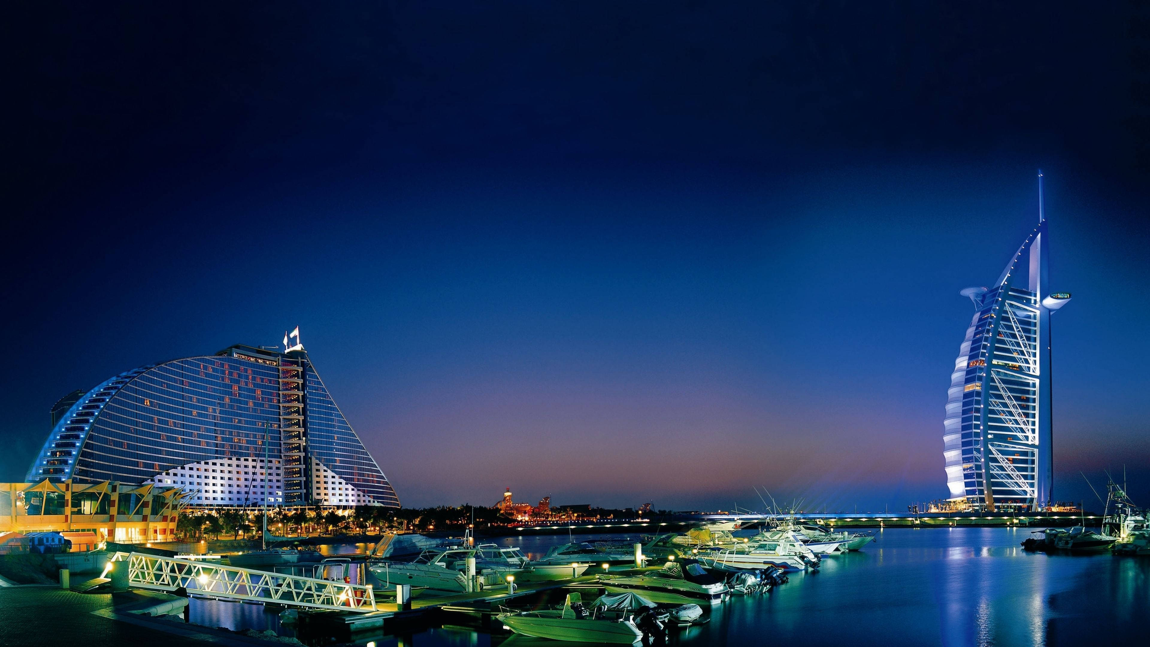 迪拜4K夜景建築 壁紙 — 杜拜標誌性帆船酒店及亞特蘭蒂斯酒店在平靜水域中倒映，並有船隻停泊的迷人夜景。