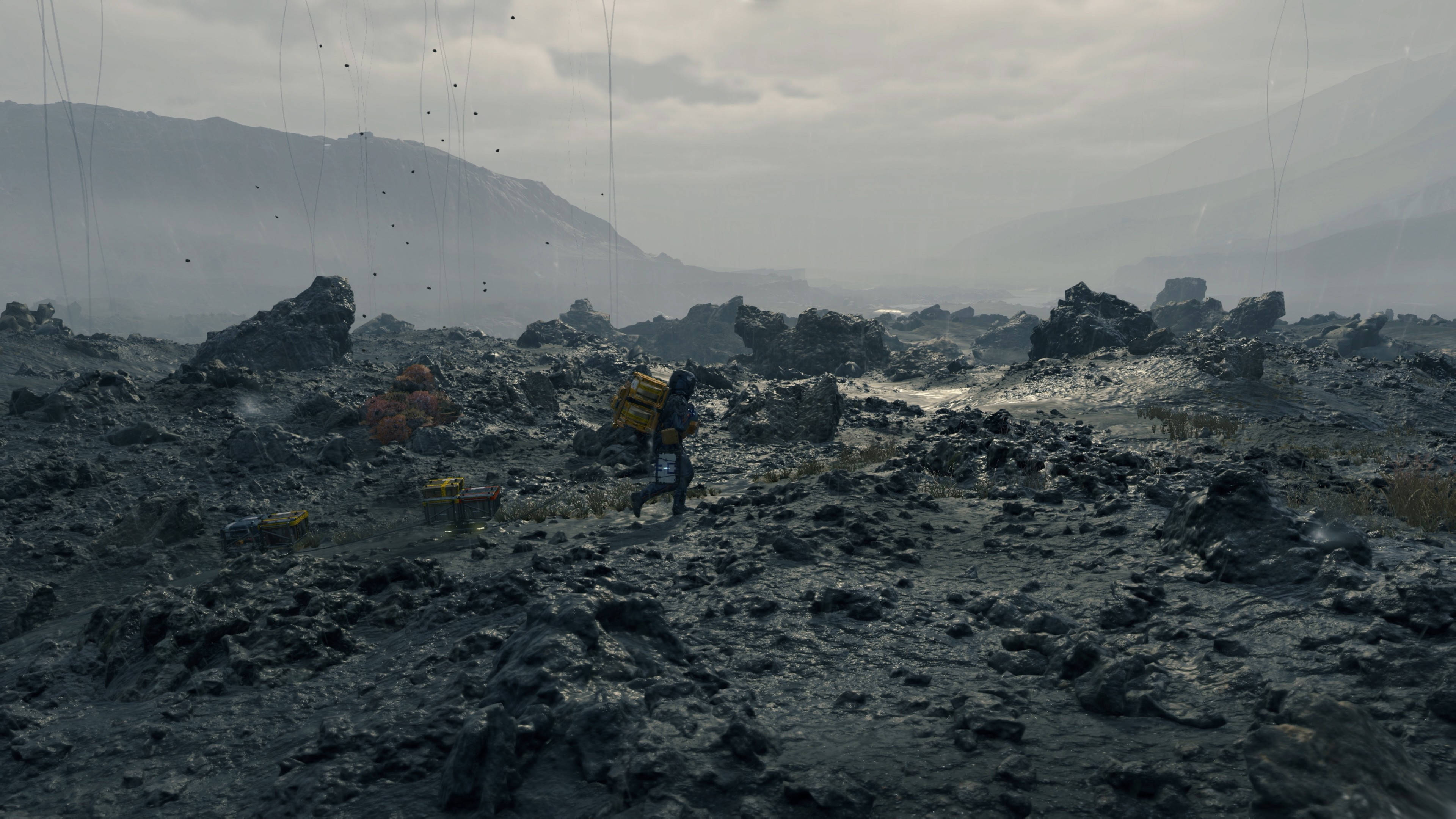 山姆行於岩石地形《死亡擱淺》4K 壁紙 — 一個孤獨的身影背著黃色背包，在陰霾密佈的天空下穿越荒涼的岩石荒地。
