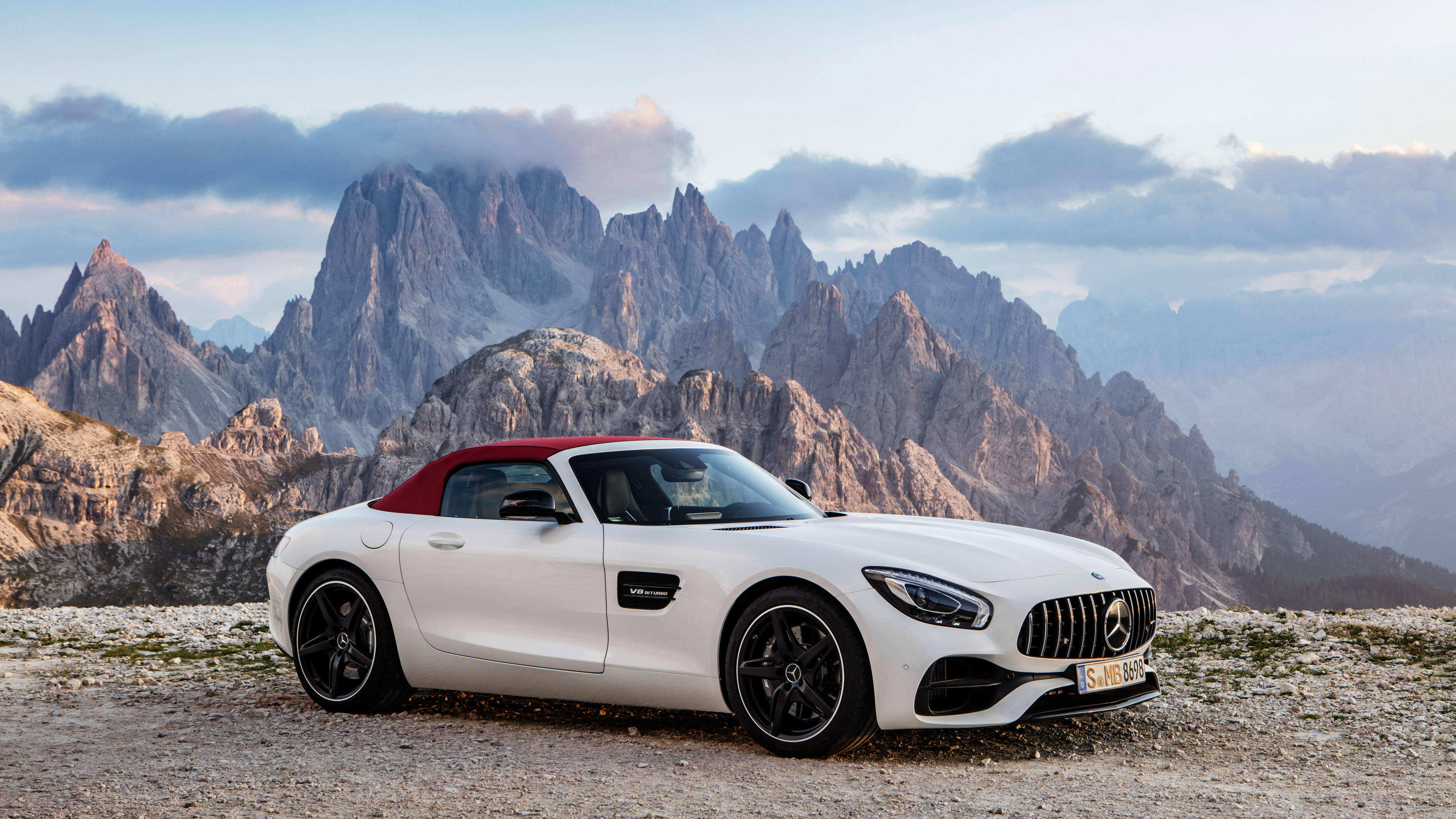 4K 奔馳敞篷白色 壁紙 — 一輛白色梅賽德斯-AMG GT Roadster，紅色軟頂，停泊在多岩石的山口，背景是鋸齒狀山峰，天空多雲。