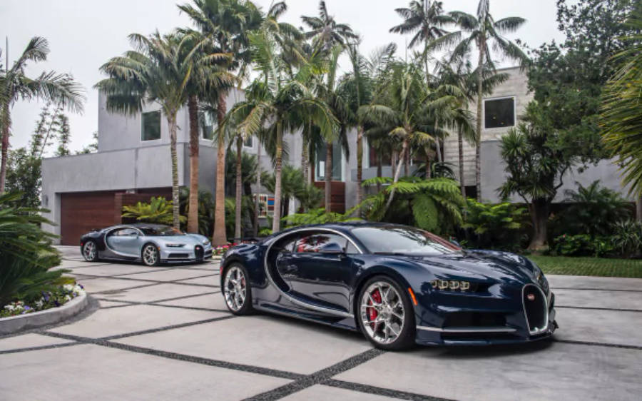 兩輛布加迪奇隆 4K 壁紙 — 兩輛時尚的Bugatti超級跑車停泊在瓷磚車道上，周圍環繞著棕櫚樹和現代豪宅。