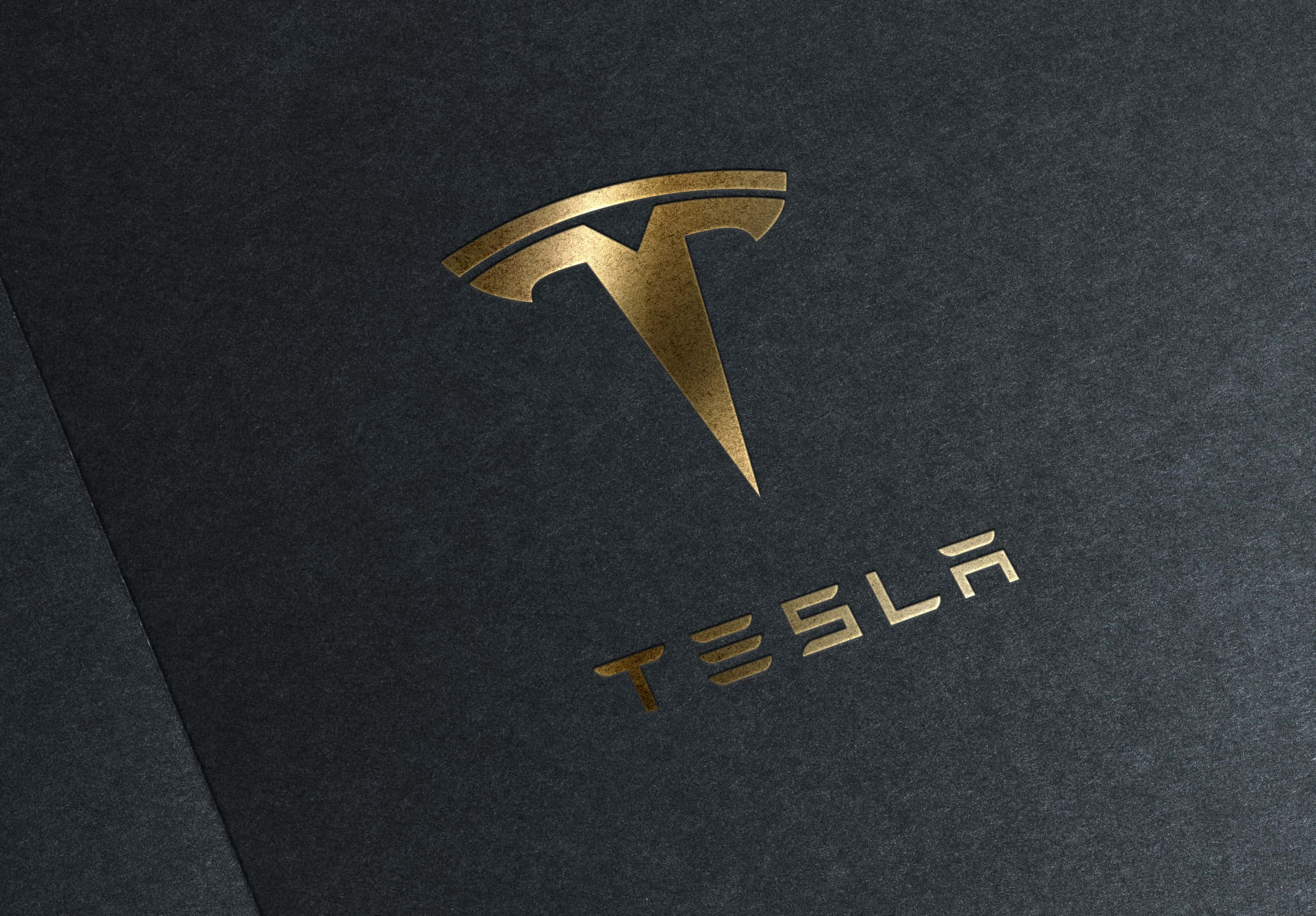 特斯拉標誌 4K 壁紙 — 金色 Tesla 標誌和文字壓印在帶紋理的深灰色紙張上，光線柔和。