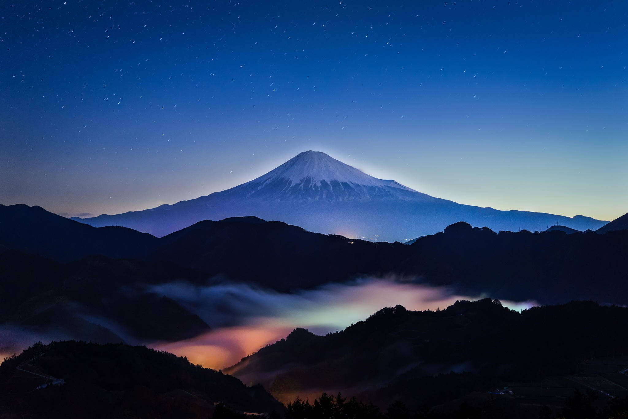 富士山日本4K夜景 壁紙 — 白雪覆蓋的富士山在繁星點點的暮色天空下雄偉地升起，下方有發光的雲彩。