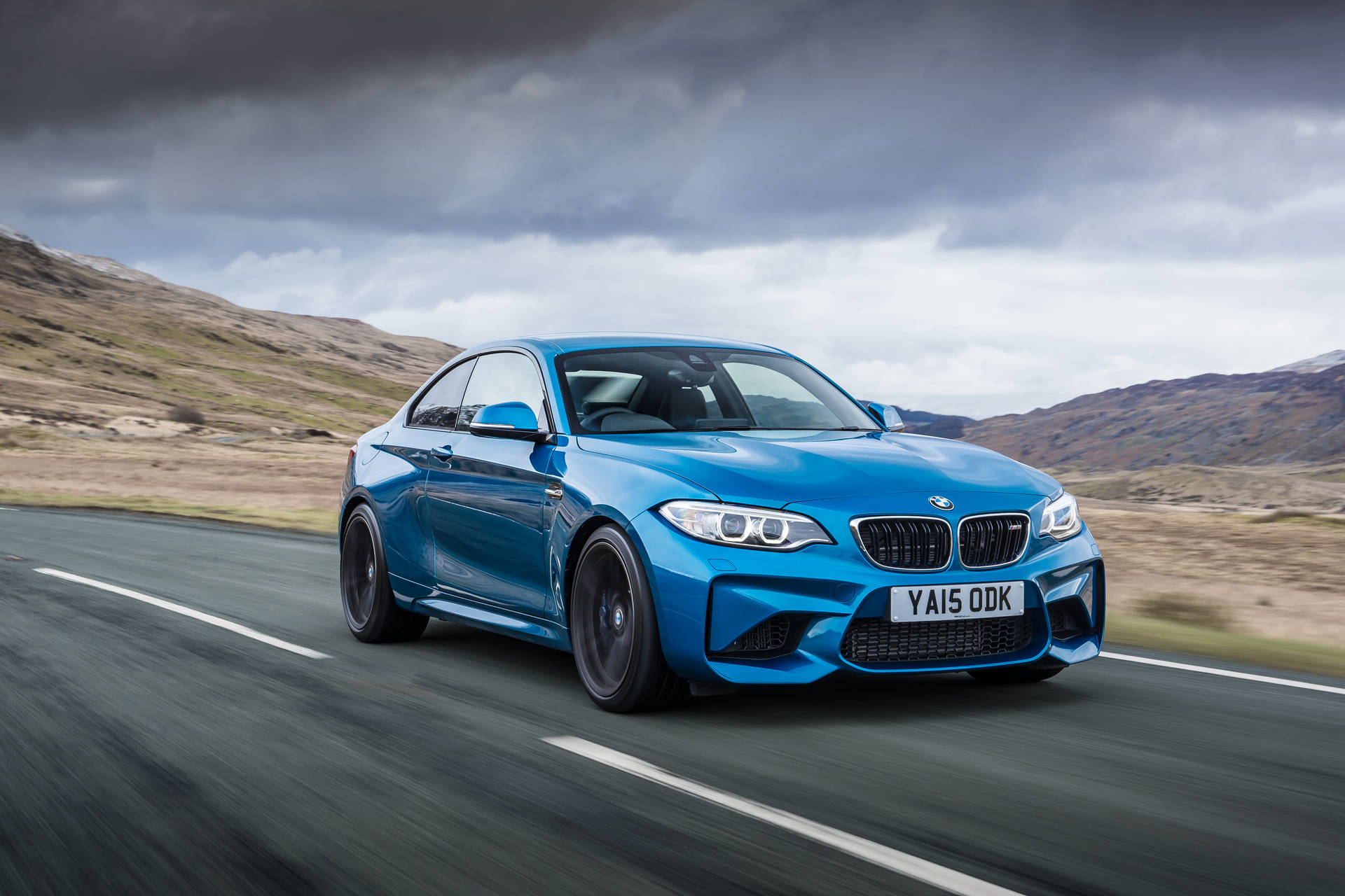 藍色BMW M2雙門跑車在多雲天空下於山路疾馳。 — 4K BMW 壁紙 壁紙
