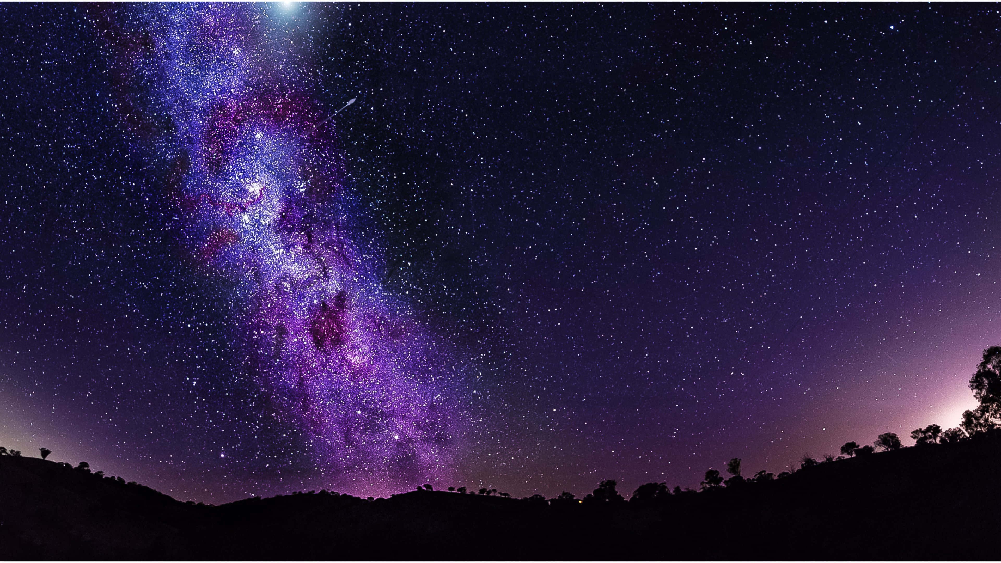 4K紫空銀河系 壁紙 — 在繁星點點的夜空下，一道壯麗的銀河在黑暗的山丘剪影上方閃耀著鮮豔的紫色和藍色色調。