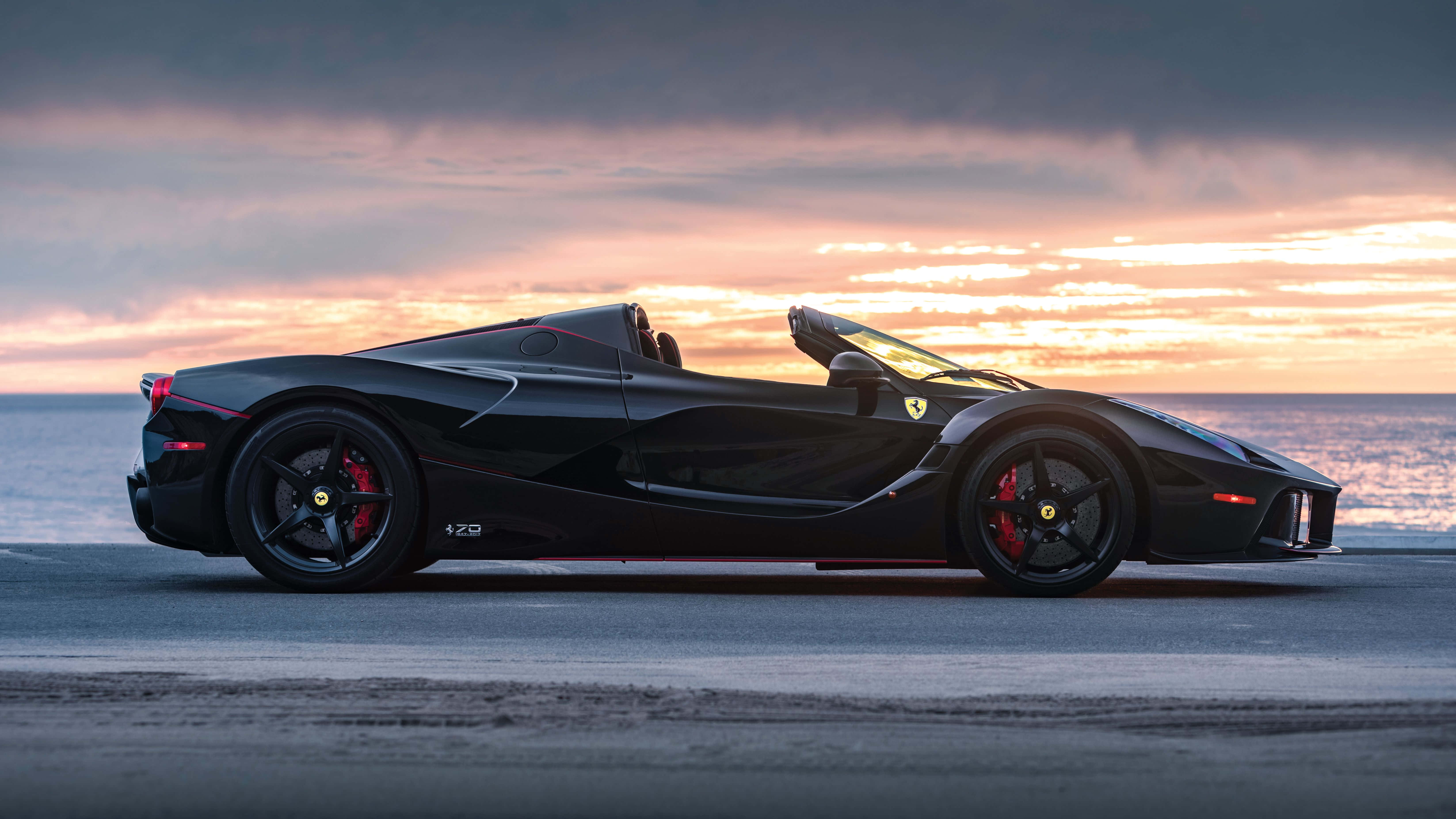 黑色法拉利跑車日落海灘4K壁紙 壁紙 — 一架時尚的黑色法拉利 LaFerrari Aperta 停泊在日落時分的海岸公路上，上空有鮮豔的橙色和粉紅色雲彩。
