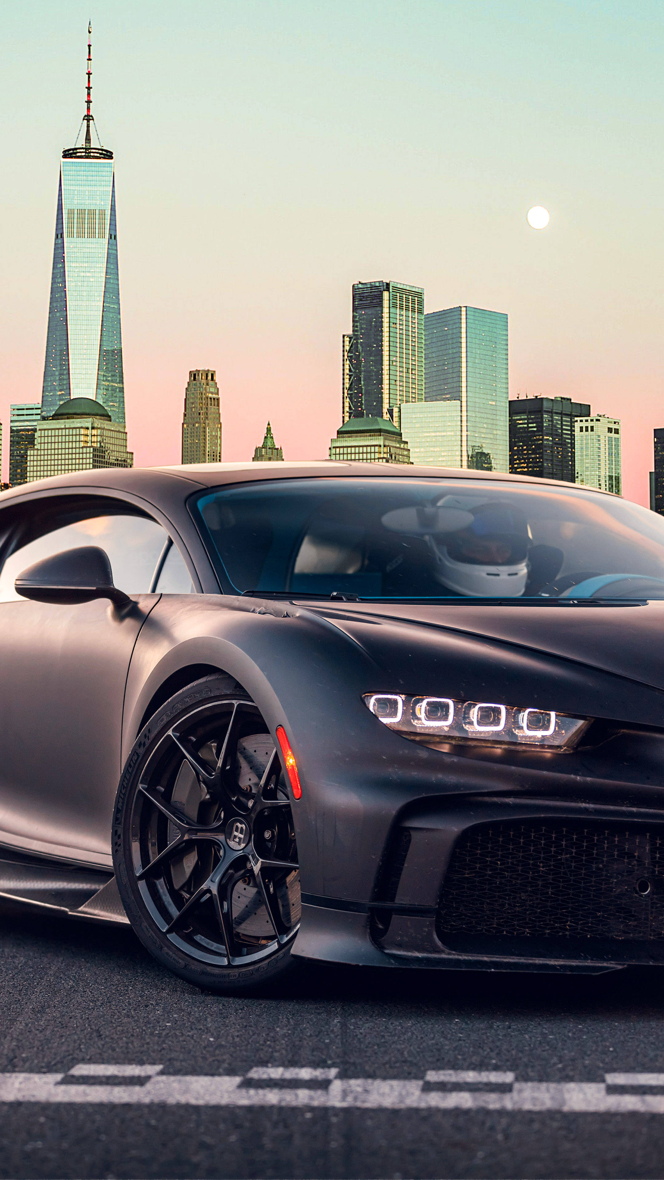 城市背景黑色 4K 車 iPhone 壁紙 — 一輛啞光黑色的Bugatti Chiron停泊在城市街道上，背景是自由塔和暮色中的月亮。