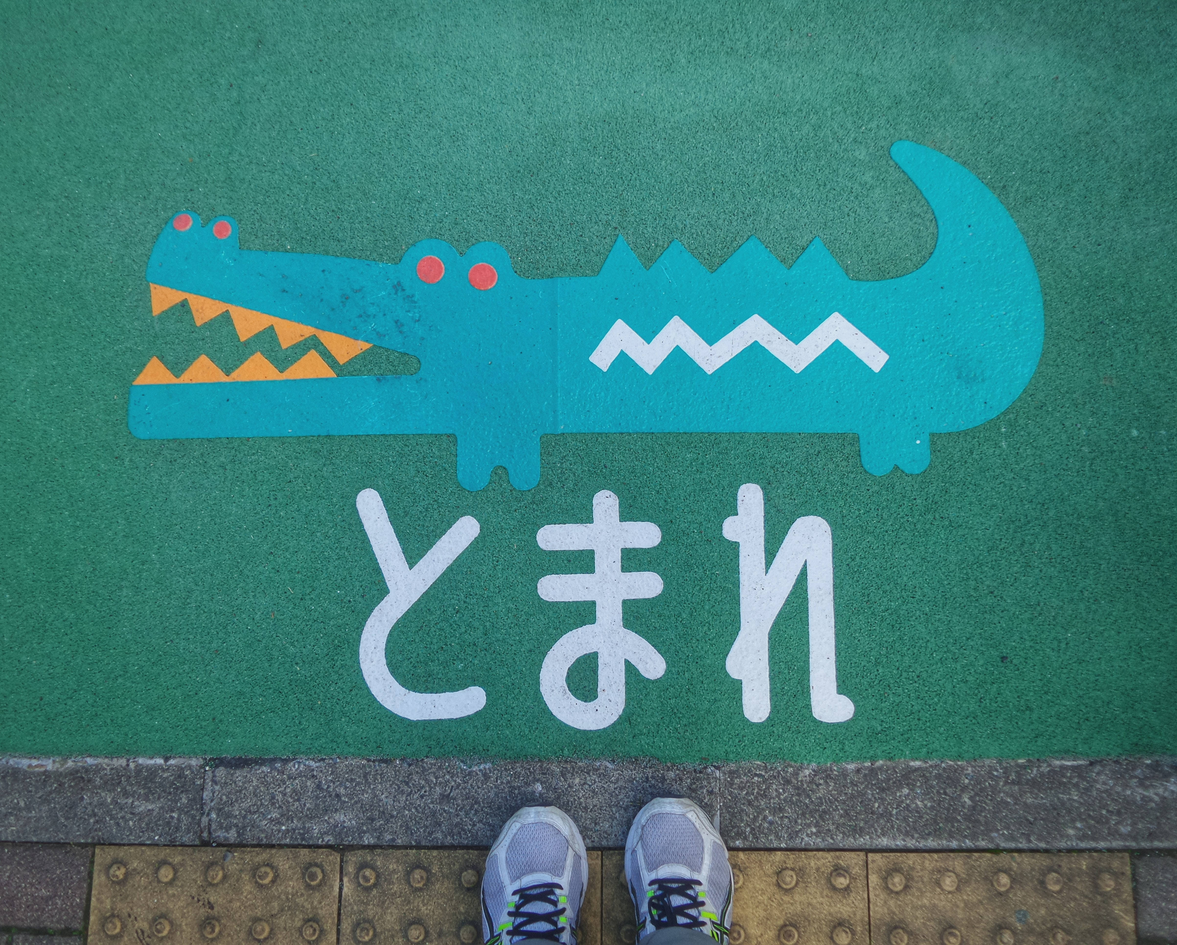 藍色鱷魚4K桌面壁紙 壁紙 — 一個人的運動鞋站在綠色遊樂場表面邊緣，上面有卡通鱷魚和日文字。