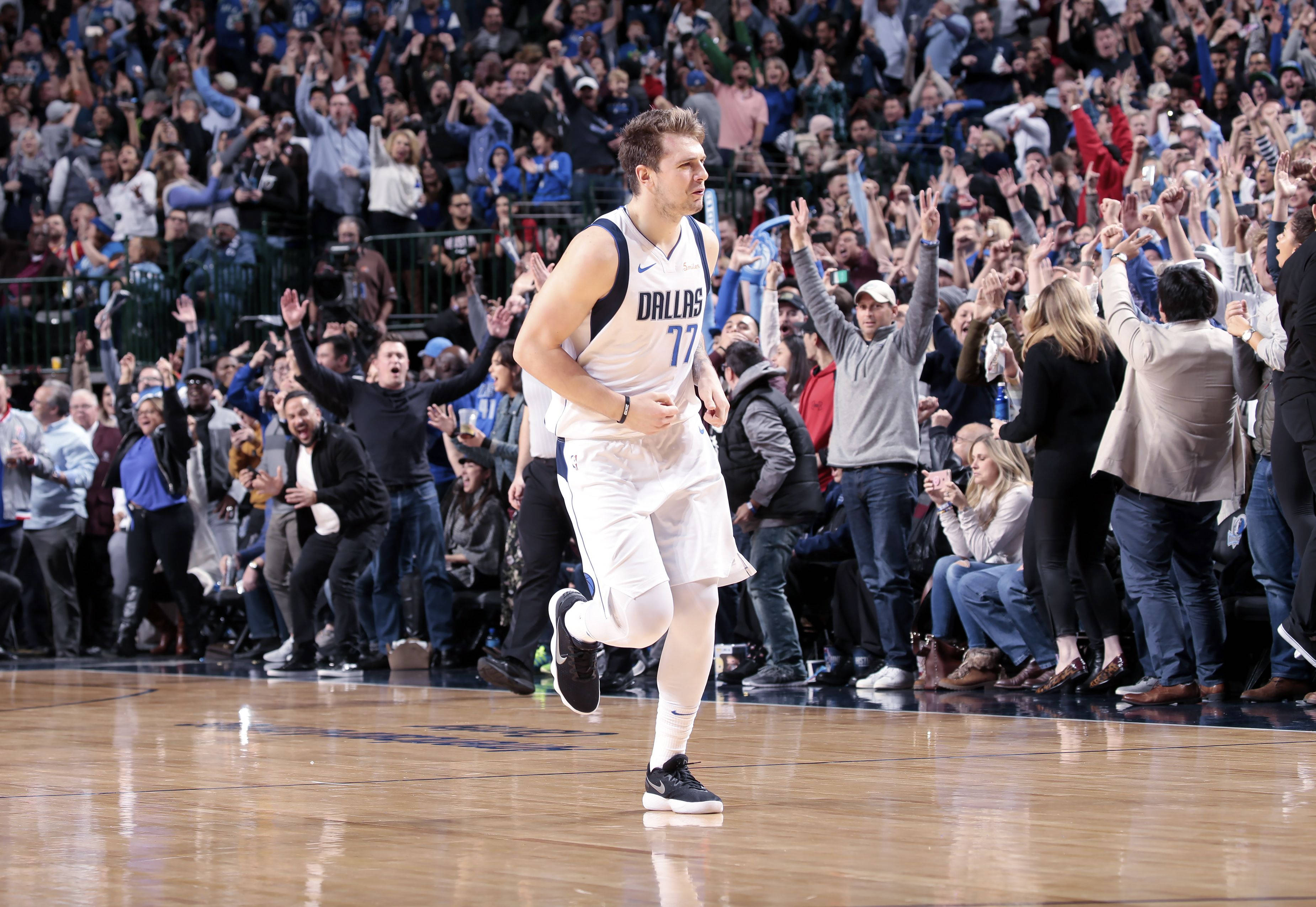 4K NBA 卢卡·多尼奇比賽瞬間 壁紙 — 一位身穿白色77號球衣的達拉斯獨行俠籃球運動員在球場上歡快地奔跑，而咆哮的球迷們歡呼並舉起雙手。