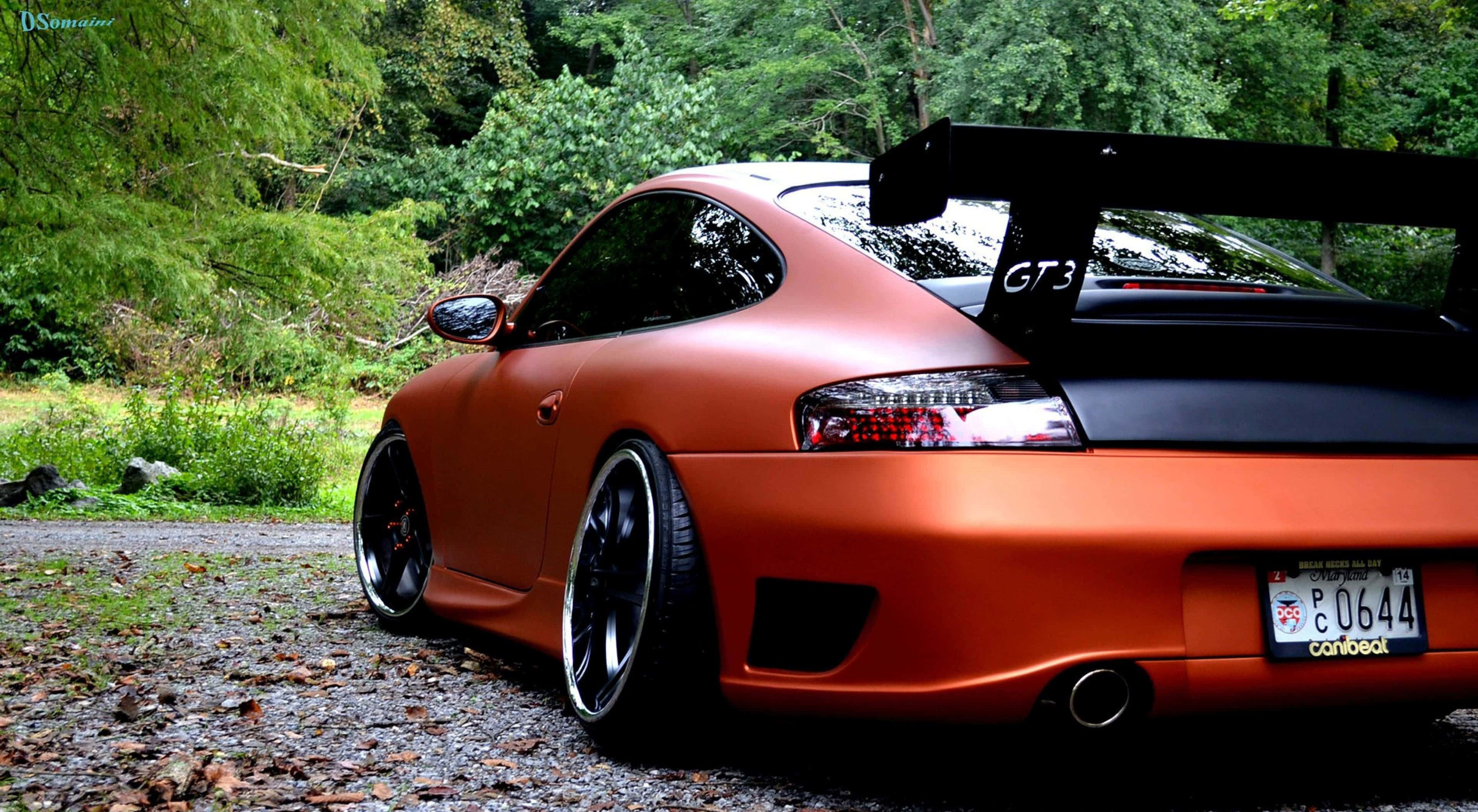 4K Project Cars 橙色 Porsche 911 背景 壁紙 — 一輛啞光橙色保時捷GT3跑車，帶有黑色飾邊，停泊在碎石路上，周圍是茂密的綠色森林。